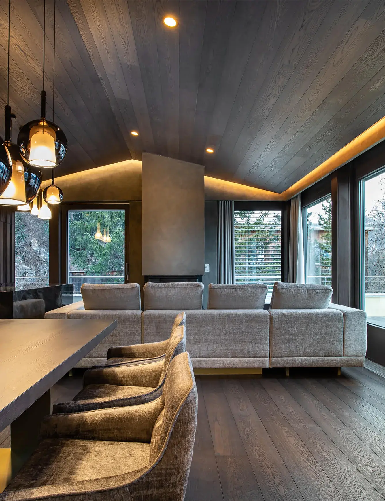Luxusní tmavý interiér Rezidence ve Svatém Mořici s kouřovou dubovou podlahou a navazujícím dřevěným podhledem s integrovaným LED podsvícením, kterému dominuje šedá sedací souprava, jídelní kout s polstrovanými křesly a designové skleněné lustry na pozadí horského výhledu.