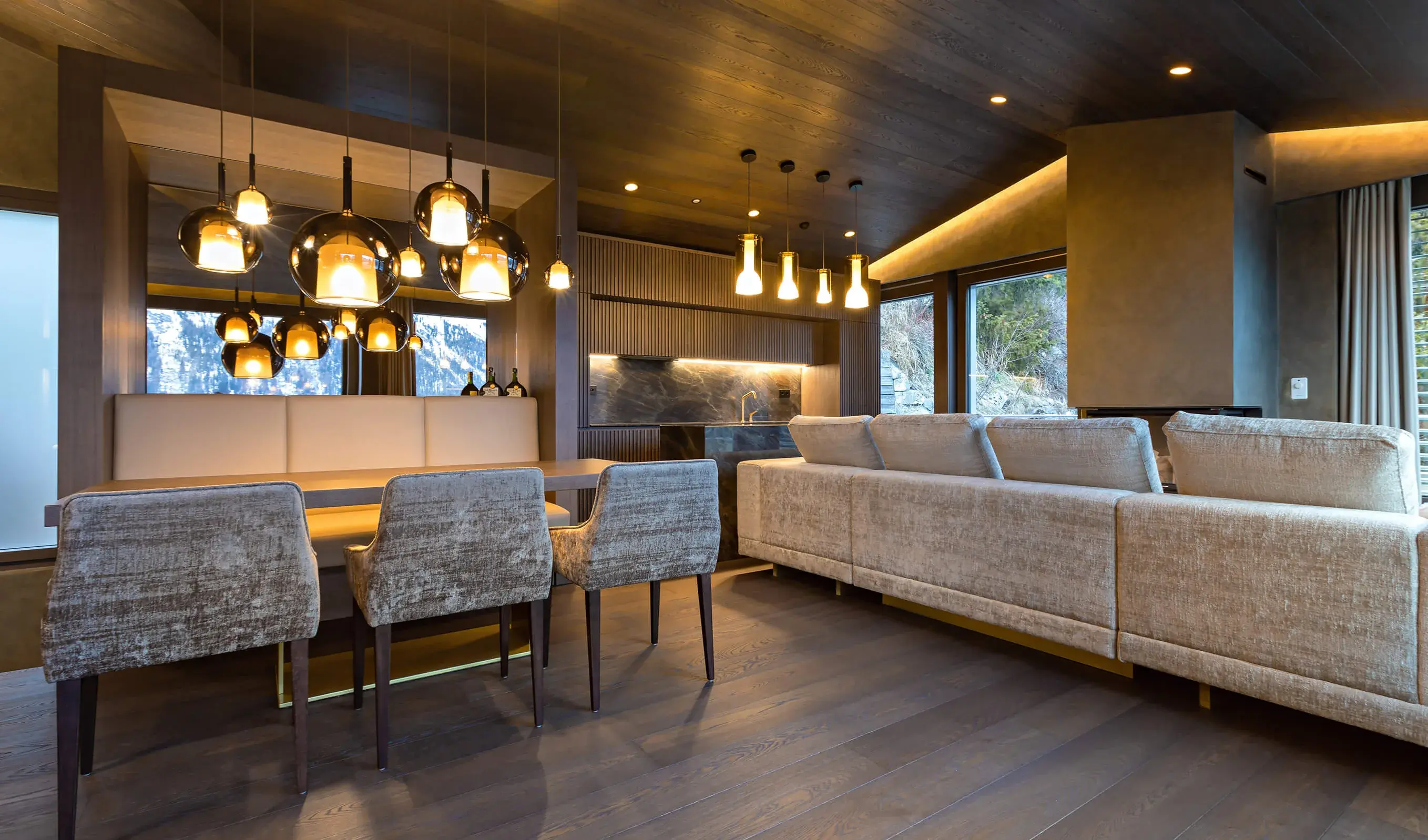 Široký záběr na hlavní obytný prostor Rezidence ve Svatém Mořici, kterému dominuje kouřová dubová podlaha Oakcent z kolekce Hazel, jejíž sytý ořechový odstín ladí s dřevěným podhledem a tmavým lamelovým obložením stěn, doplněný o světlou modulární pohovku a jídelní kout s luxusními polstrovanými křesly.