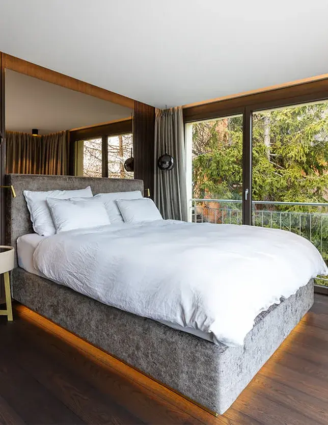 Moderní ložnice v Rezidenci ve Svatém Mořici s tmavou dubovou podlahou a velkým zrcadlem nad čelem postele, které opticky zvětšuje prostor a odráží výhled z protilehlého proskleného balkonu do okolní zeleně.