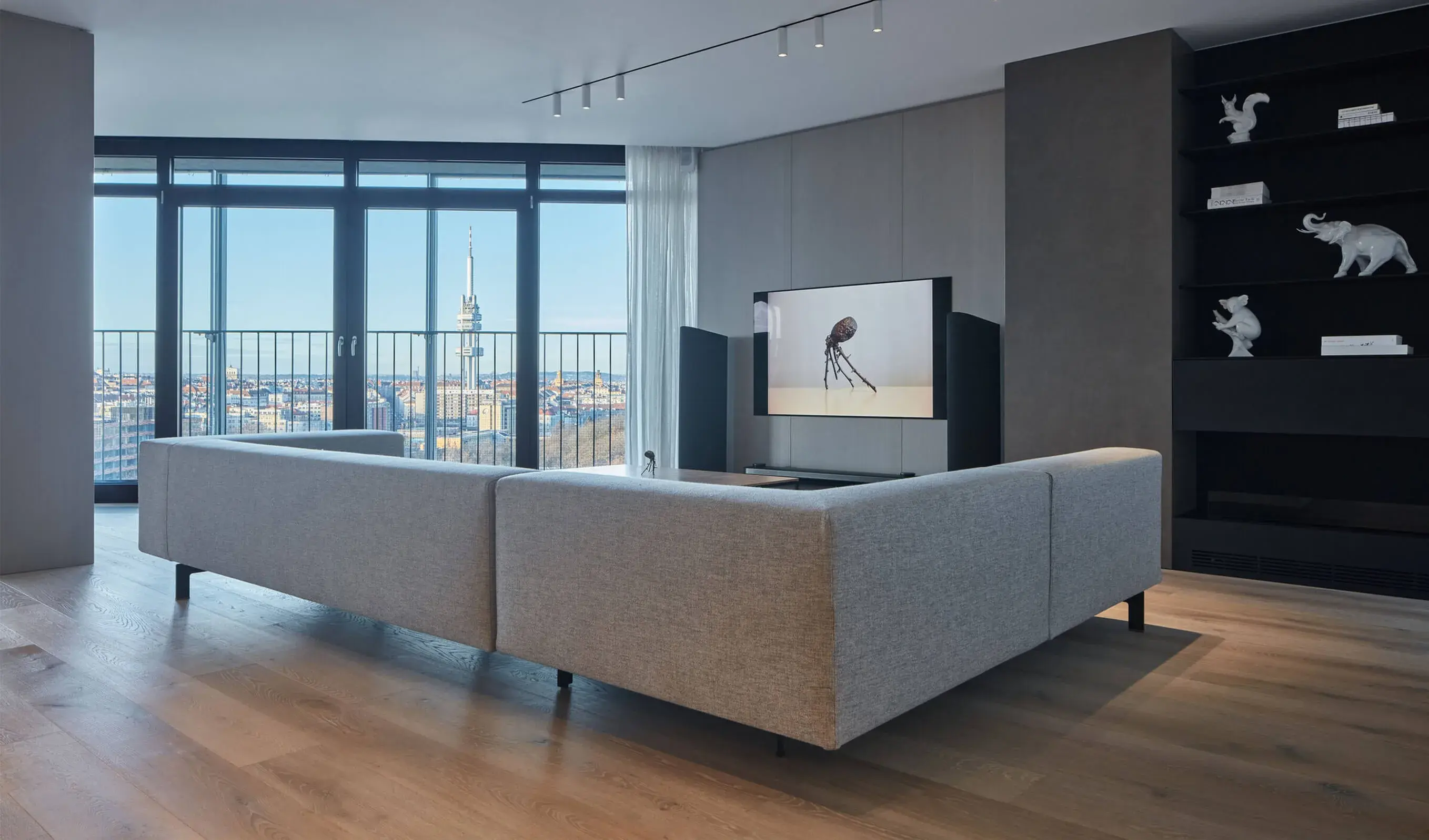 Celkový pohled na minimalistický obývací pokoj s šedou rohovou pohovkou, černou knihovnou a velkorysým prosklením, které nabízí panoramatický výhled na Prahu s dominantou Žižkovské věže nad kartáčovanou dubovou podlahou Oakcent.