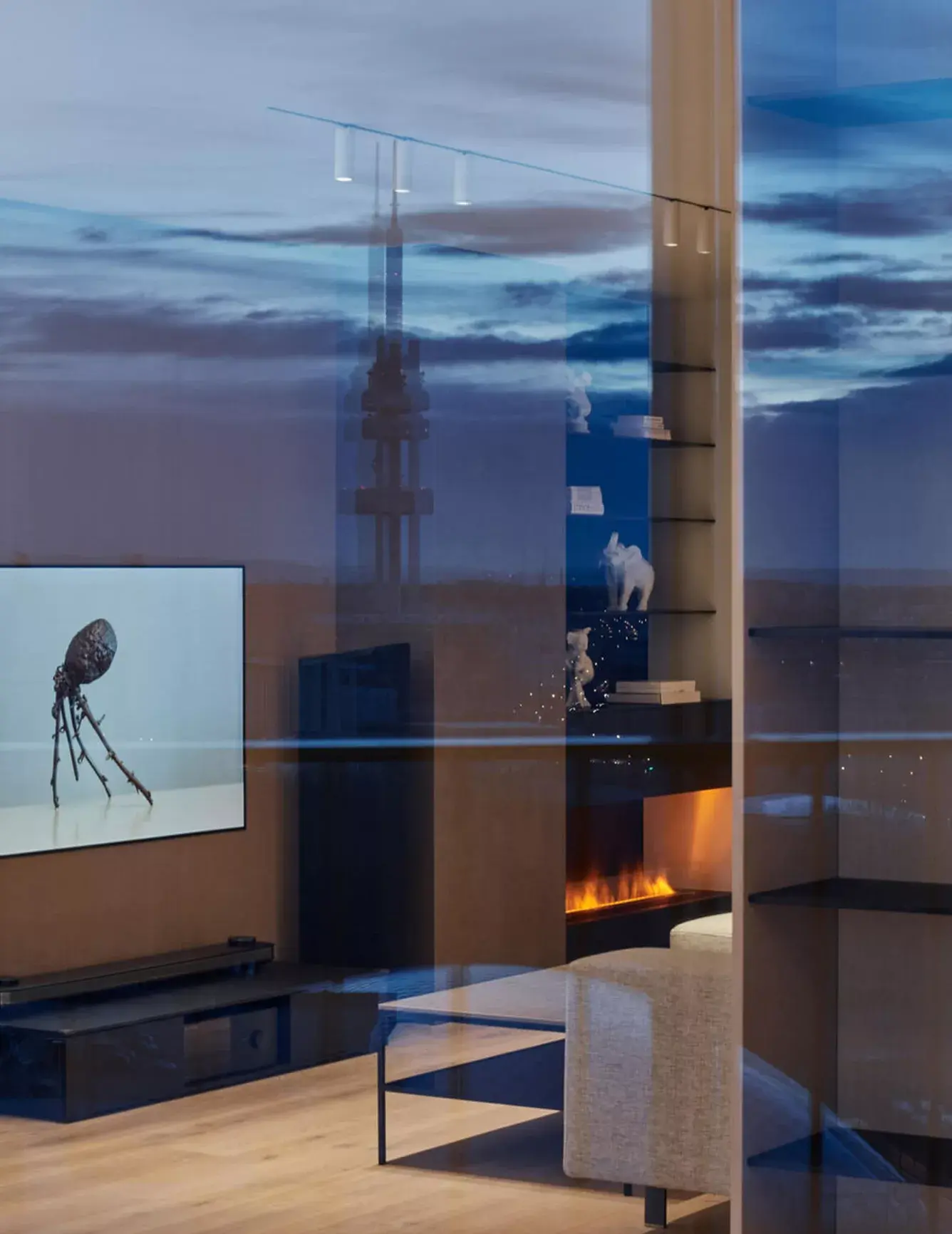 Večerní atmosféra v minimalistickém obývacím pokoji s výhledem na osvětlenou Žižkovskou věž, jejíž silueta se odráží v prosklené stěně vedle moderního krbu a dubové podlahy Oakcent.