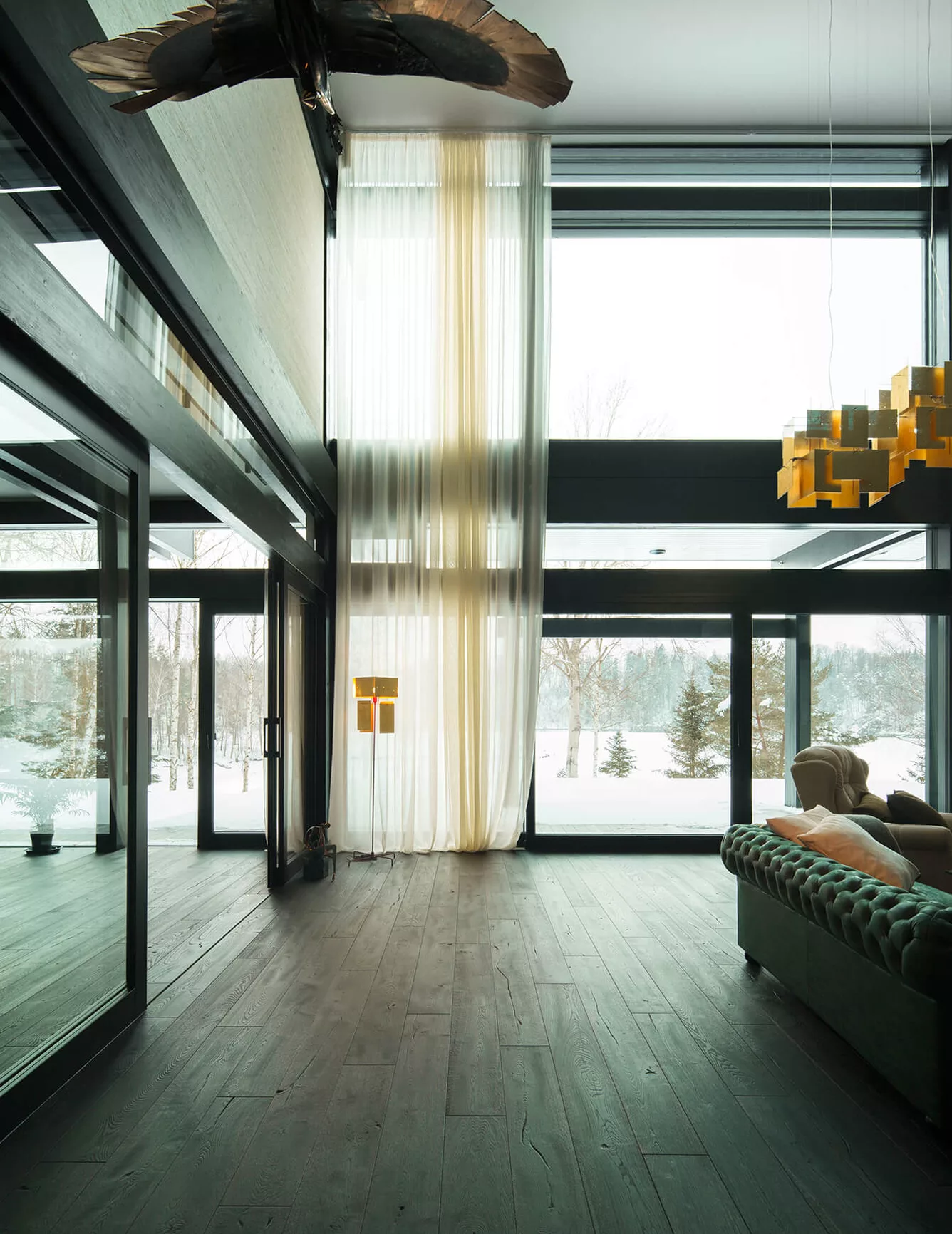 Moderní prostorný interiér s vysokými stropy a velkoformátovými okny, kterému dominuje tmavá dubová podlaha z kolekce Ink v kontrastu se světlými závěsy a výhledem do zasněžené zahrady.