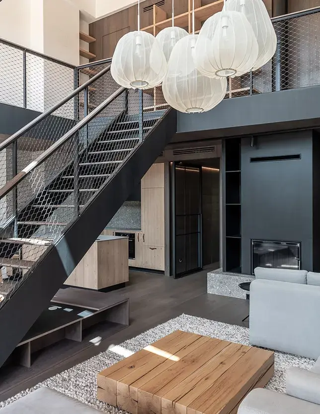 Celkový pohled na mezonetový obývací pokoj v apartmánu Lipno s šedou dubovou podlahou z kolekce Smoke, černým ocelovým schodištěm, krbem a výrazným designovým osvětlením v moderním šumavském stylu.