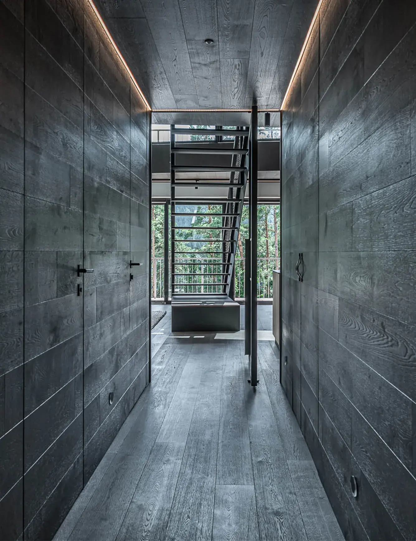 Pohled do moderní chodby apartmánu na Lipně s tmavě šedým dubovým obkladem stěn a podlahou Oakcent, která směřuje k prosklenému schodišti s výhledem do lesa.