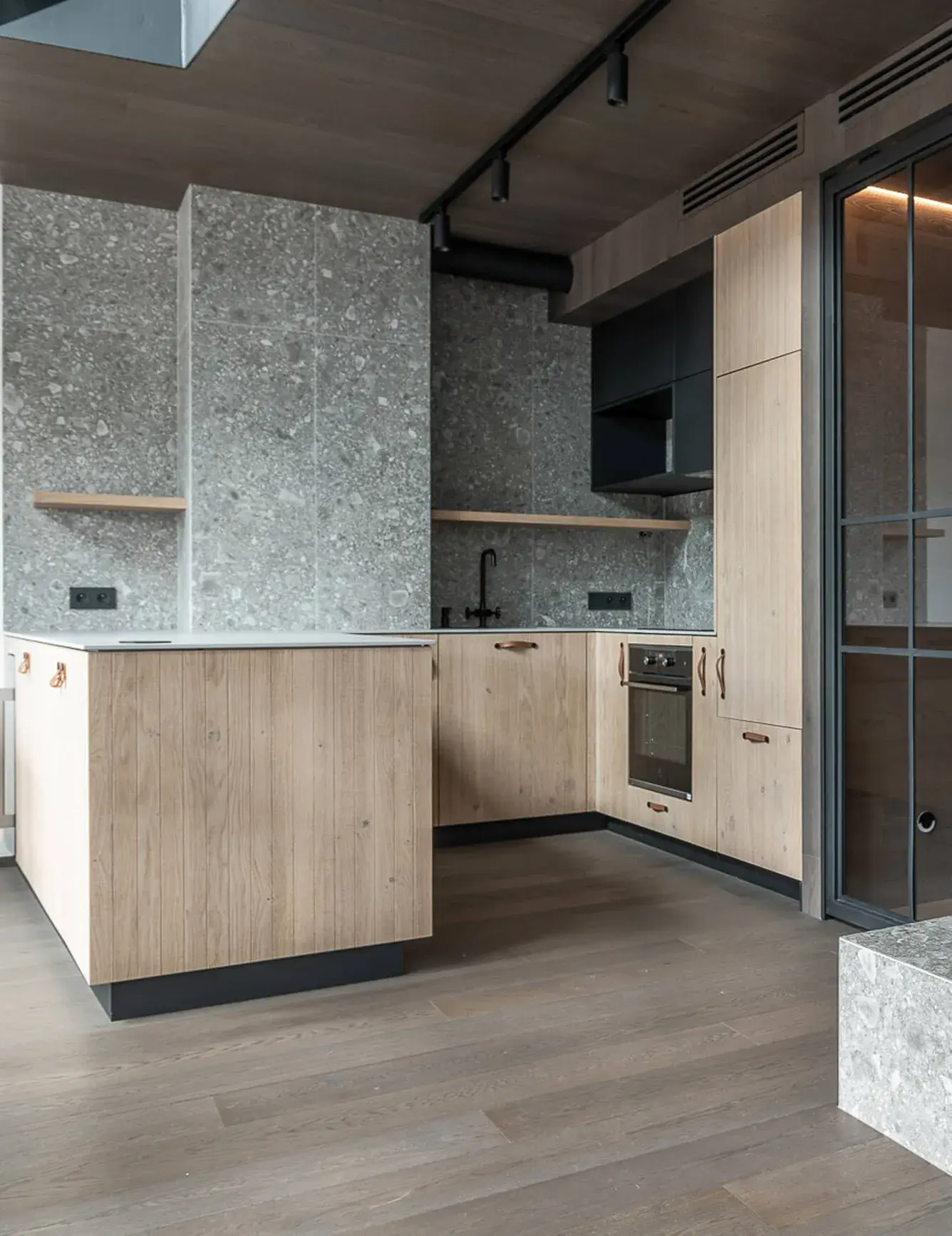 Pohled na minimalistickou kuchyni v apartmánu Lipno s dubovým kuchyňským ostrovem a linkou Oakcent, šedým terazzo obkladem stěn a tmavou dřevěnou podlahou, doplněný o černé industriální osvětlení.
