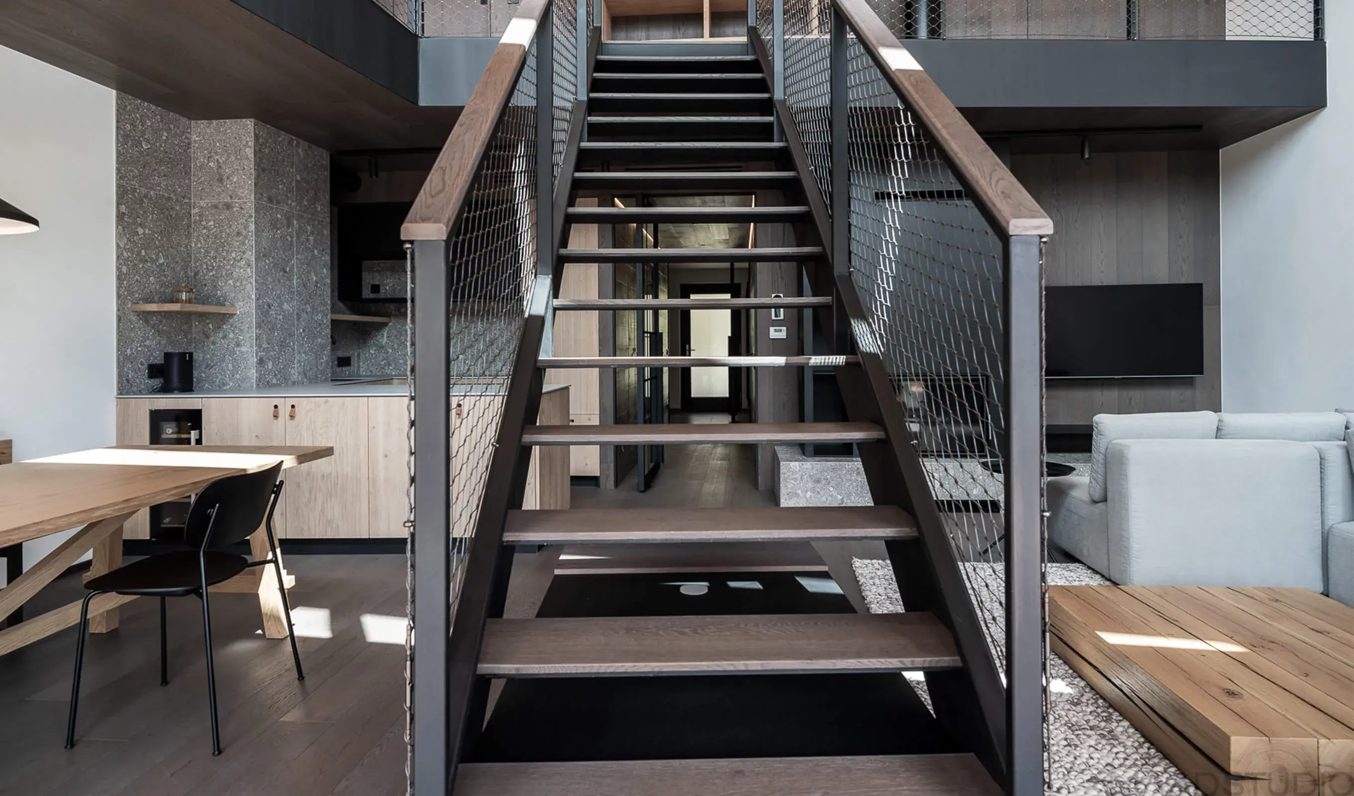 Pohled skrze černé schodiště s dřevěnými nášlapy na otevřený interiér apartmánu kombinující dub Oakcent, kámen a industriální prvky.
