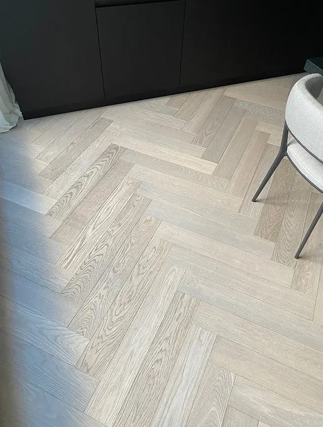 Detailní pohled na dubovou podlahu Oakcent Premium položenou ve vzoru Herringbone (klasický stromeček) v odstínu Milky, která plynule navazuje na černou soklovou lištu minimalistické kuchyňské linky v apartmánu na Josefově.