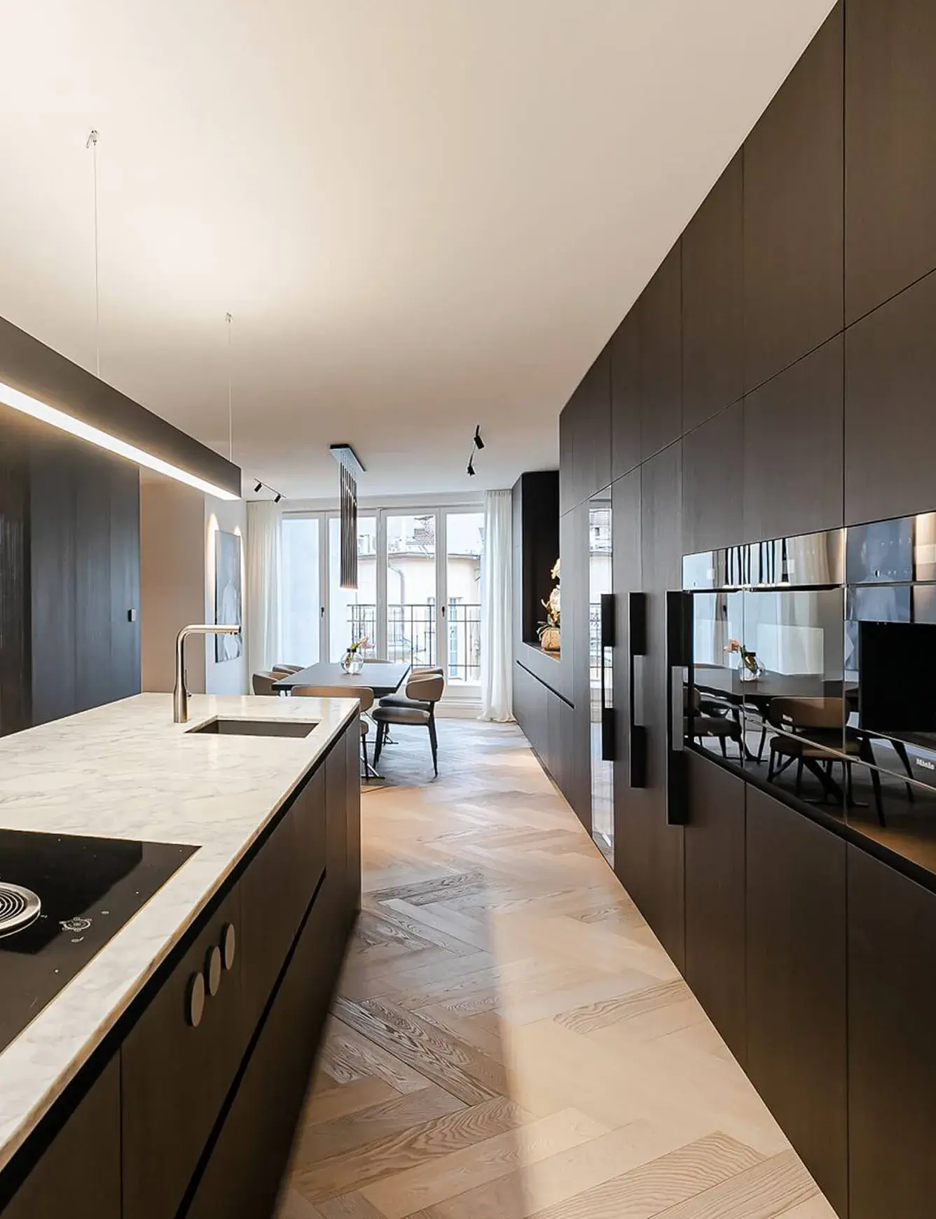Široký průhled do interiéru soukromého bytu na pražském Josefově, kde dubová podlaha Oakcent Premium v klasickém vzoru Herringbone (stromeček) opticky propojuje moderní černou kuchyni s mramorovou deskou a jídelní kout, prosvětlený velkými okny v pozadí.