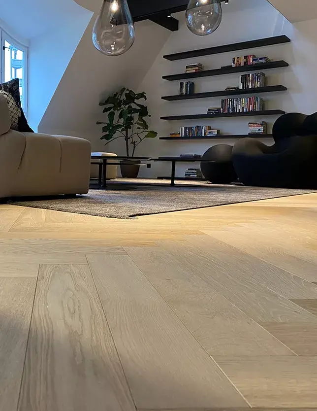 Detailní pohled na světlou dubovou podlahu Oakcent Premium ve vzoru Herringbone (stromeček), která plynule přechází podél designové sedací soupravy k černé otevřené knihovně v podkrovním apartmánu na Josefově.