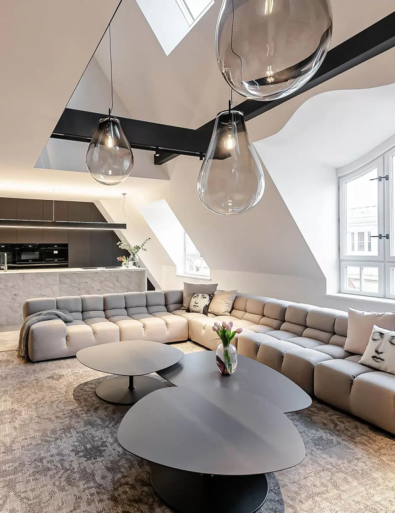 Široký pohled do podkrovního obývacího pokoje apartmánu na Josefově, kterému dominuje velká modulární sedací souprava v béžové barvě, designové konferenční stolky a výrazná skleněná svítidla ve tvaru kapek zavěšená na přiznaných černých trámech.