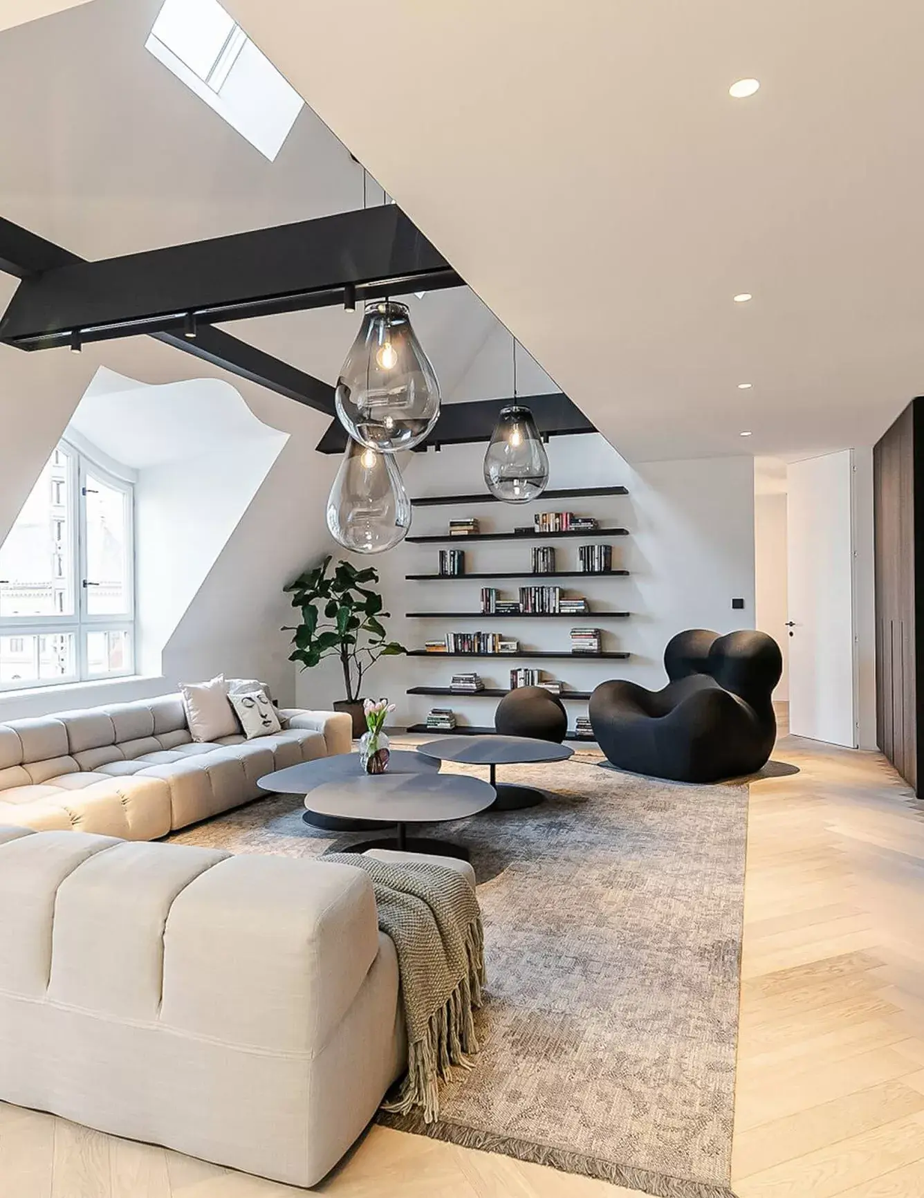 Alt: Široký pohled do prosvětleného obývacího pokoje v podkrovním apartmánu na Josefově, kde světlou podlahu Oakcent Premium Herringbone v odstínu Milky doplňuje velká modulární pohovka v krémové barvě a designové černé doplňky pod přiznanými trámy.