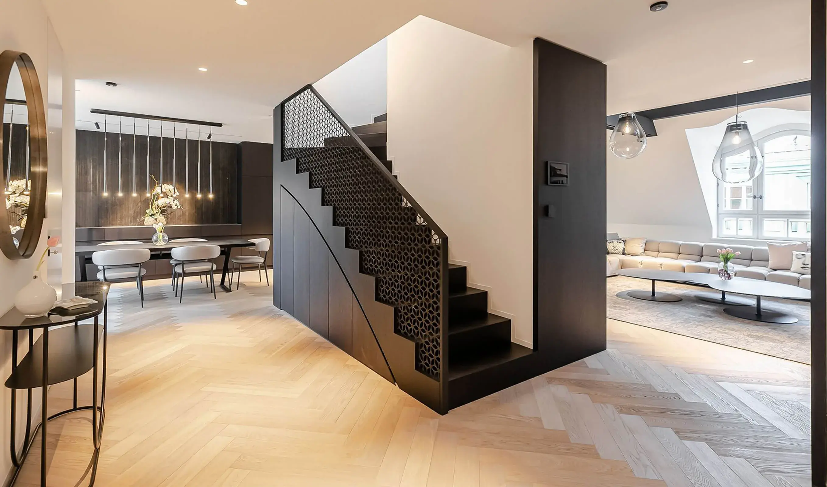 Široký pohled na prosvětlený interiér pražského apartmánu na Josefově, kde světlou podlahu Oakcent Premium Herringbone v odstínu Milky doplňuje výrazné černé schodiště s dekorativním perforovaným zábradlím a designová jídelna s mramorovým stol