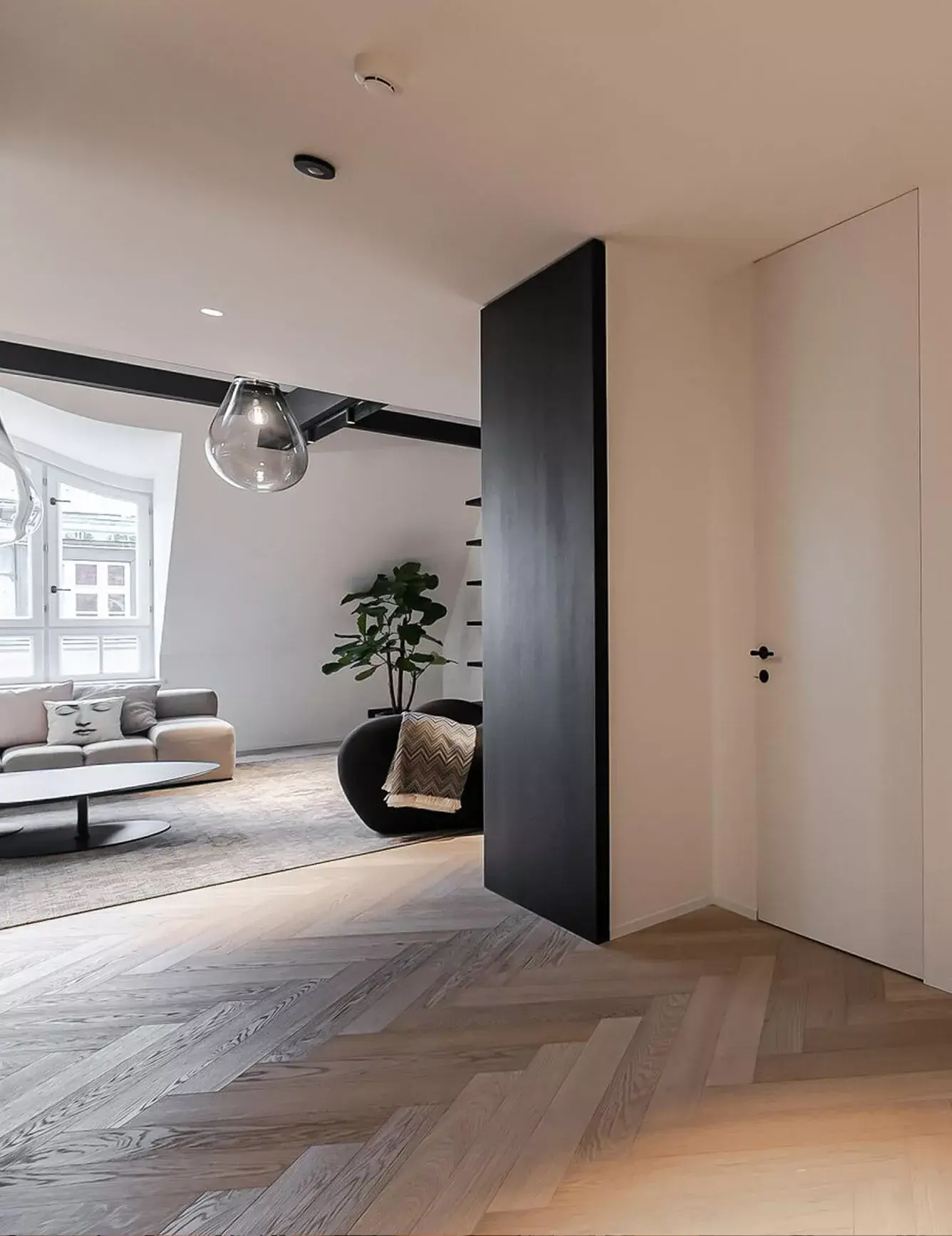 Pohled z předsíně do obývacího pokoje apartmánu na Josefově, kde dubová podlaha Oakcent Premium Herringbone v odstínu Milky plynule přechází pod černé designové dveře a otevírá prostor k prosvětlenému interiéru s velkou sedací soupravou.