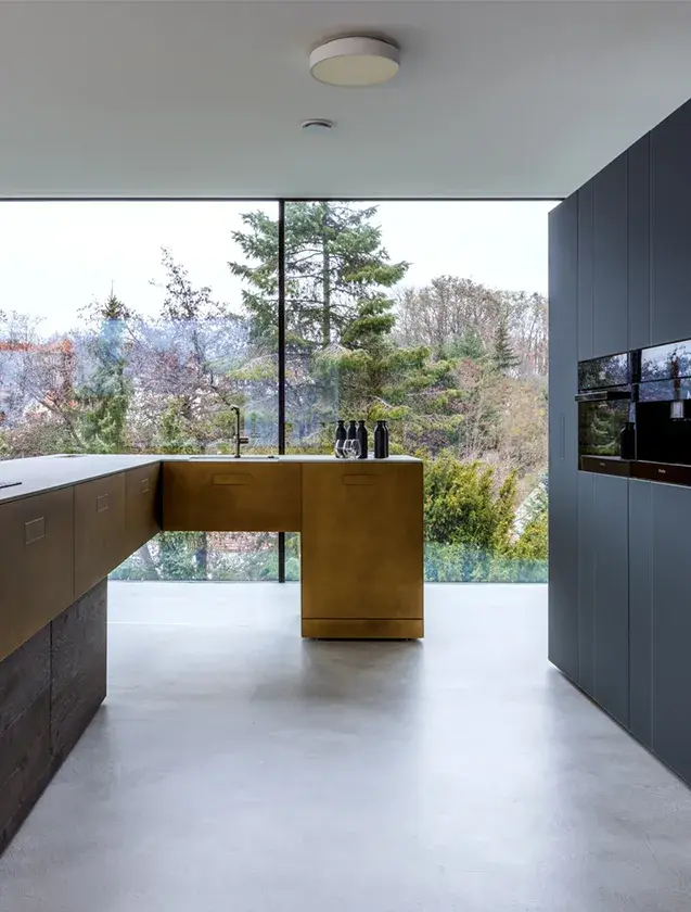 Moderní kuchyně s jídelním koutem v realizaci LAGO Showhouse, kde velkoformátové prosklení otevírá prostor do zahrady a světlá podlaha podtrhuje koncept kontinuálního interiéru