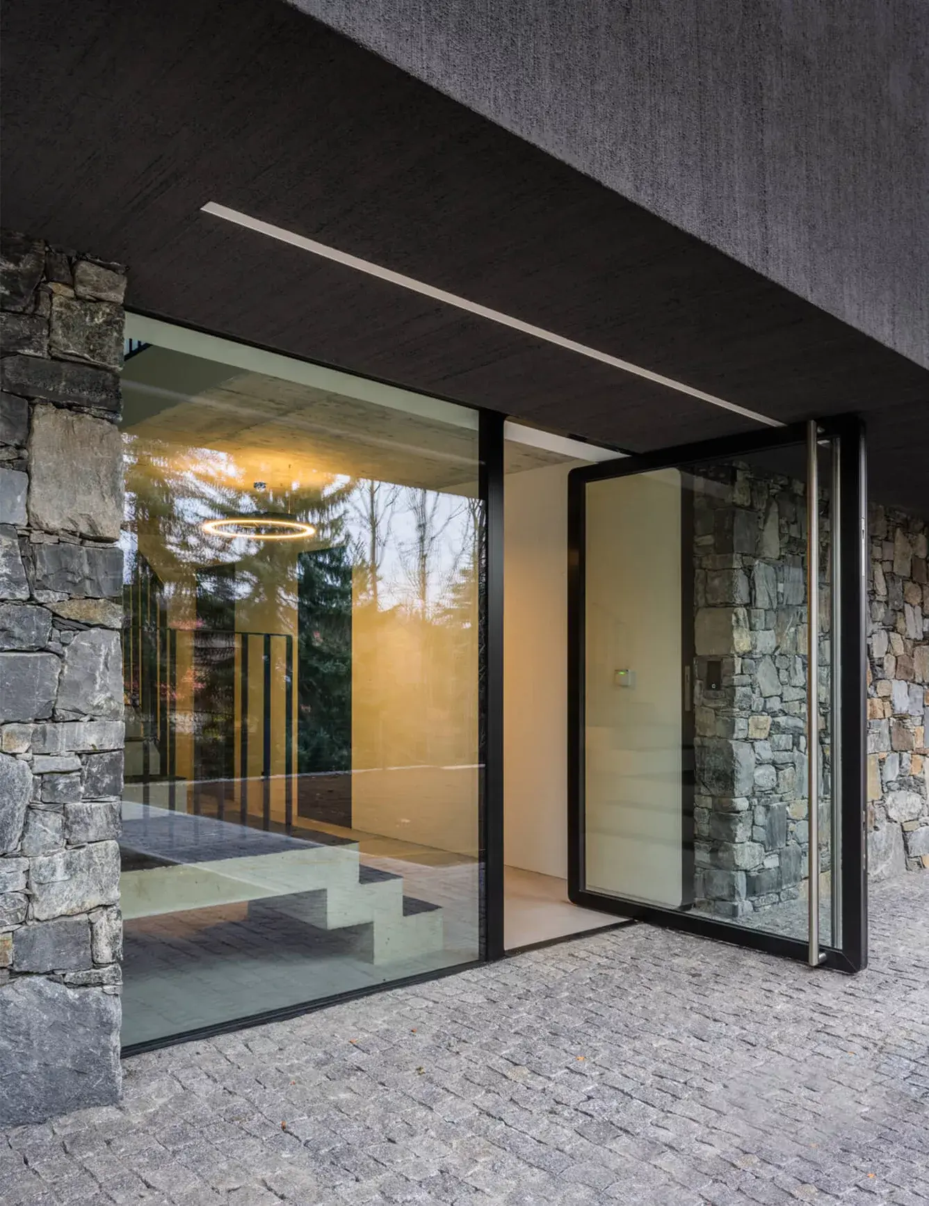 Pohled na vstup do moderní vily s fasádou z přírodního kamene a velkoformátovými skleněnými dveřmi, skrze které prosvítá minimalistický interiér s designovým osvětlením.