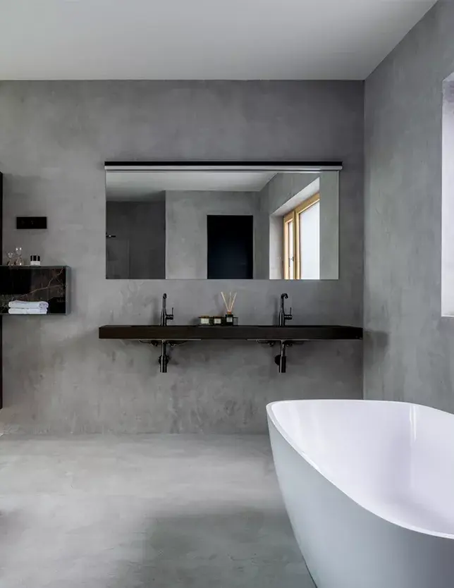 Moderní minimalistická koupelna v realizaci LAGO Showhouse s šedou stěrkou na stěnách i podlaze, které dominuje volně stojící bílá vana a tmavá zavěšená deska se dvěma umyvadly pod velkým zrcadlem.