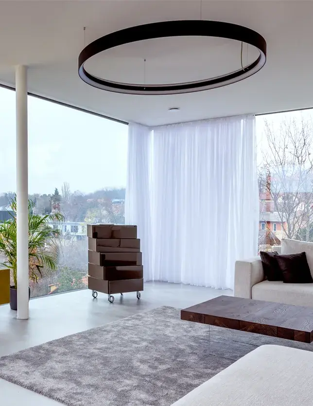 Prosvětlený obývací pokoj v realizaci LAGO Showhouse s velkoformátovým rohovým prosklením, kterému dominuje kruhové závěsné svítidlo, minimalistická komoda na kolečkách a dřevěný konferenční stolek na skleněné podnoži.