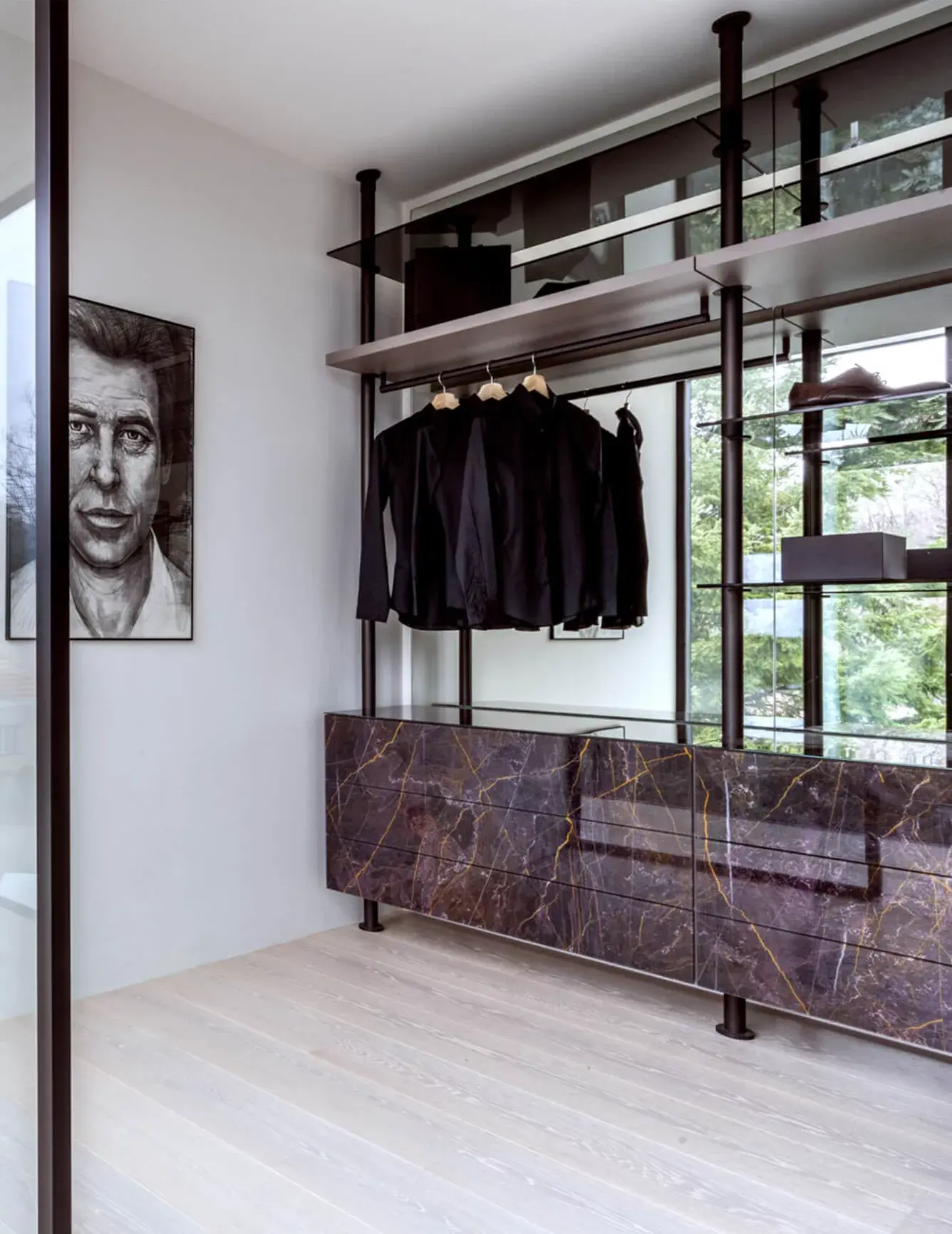 Moderní šatna v realizaci LAGO Showhouse s otevřeným policovým systémem a komodou v dekoru tmavého mramoru, doplněná světlou dřevěnou podlahou a uměleckým portrétem na stěně.