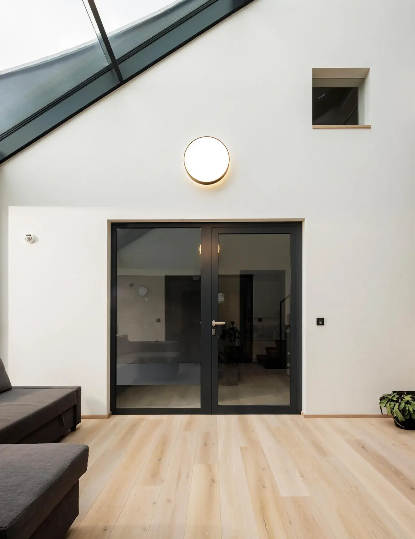 Interiér moderního obývacího pokoje v Židlochovicích s proskleným atriem, kde světlá dubová podlaha OAKCENT ladí s bílou stěnou s minimalistickým kruhovým svítidlem a kontrastní černou pohovkou pod zkoseným stropem se střešním oknem.