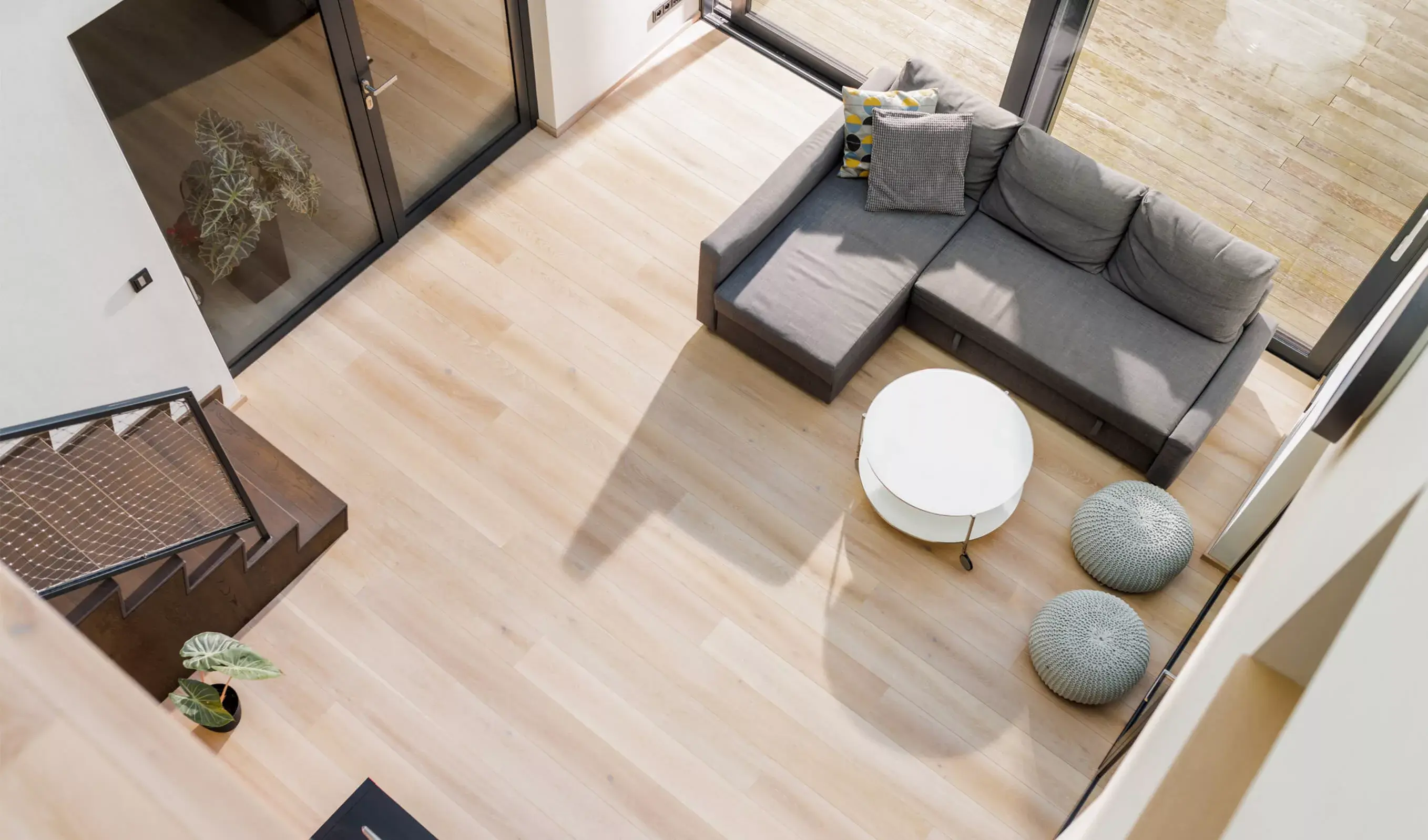 Pohled z ptačí perspektivy do prosvětleného obývacího pokoje v Židlochovicích, kde velmi světlá dubová podlaha Oakcent Ivory tvoří čistý základ pro šedou rohovou pohovku a kontrastuje s tmavým schodištěm v kaštanově hnědém odstínu kolekce Hazel.