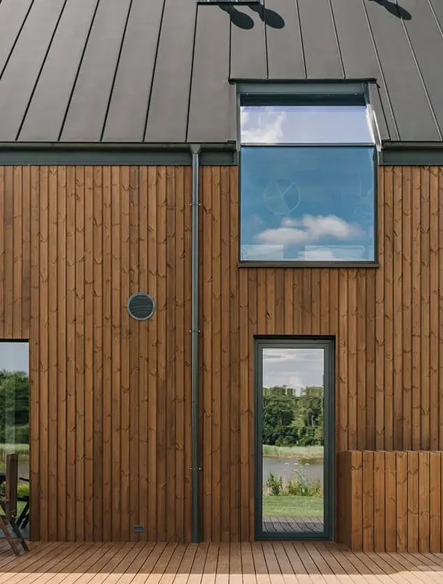 Detailní pohled na fasádu moderní stodoly v Miloslavě od Razoo architekti, kterou tvoří svislé dřevěné obklady v teplém přírodním odstínu, doplněné o antracitovou plechovou střechu s integrovaným střešním oknem a velkorysé prosklené plochy s výhledem do krajiny.