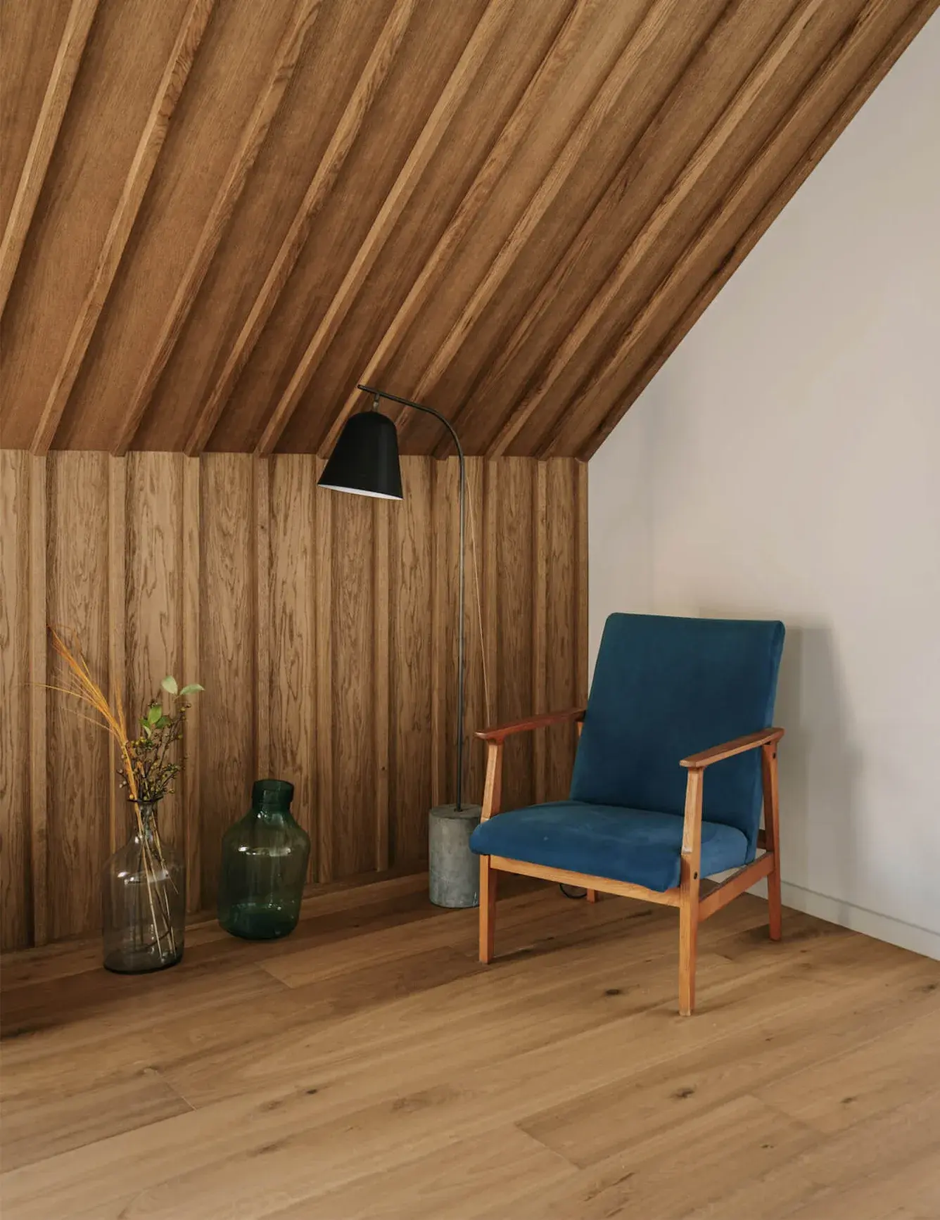 Detail koutu v podkroví stodoly v Miłoslawi, kde dubová podlaha Oakcent plynule přechází v dekorativní svislé obložení stěny a šikmý lamelový podhled, doplněný retro modrým křeslem a designovými doplňky.