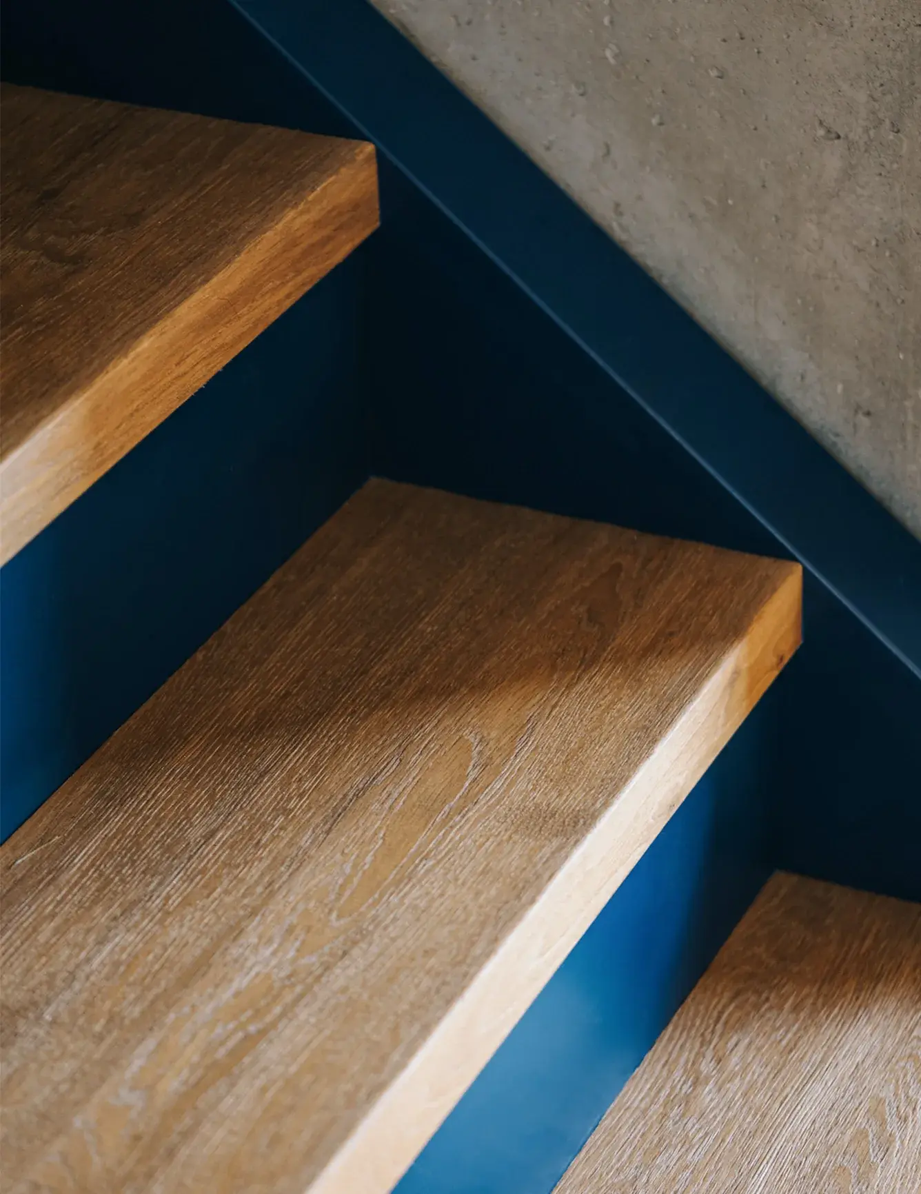 Detail schodišťového stupně z masivního dubu OAKCENT, který je zasazen do kontrastní sytě modré bočnice schodiště a doplňuje industriální estetiku interiéru s pohledovou betonovou stěnou v projektu stodoly v Miłoslawi.