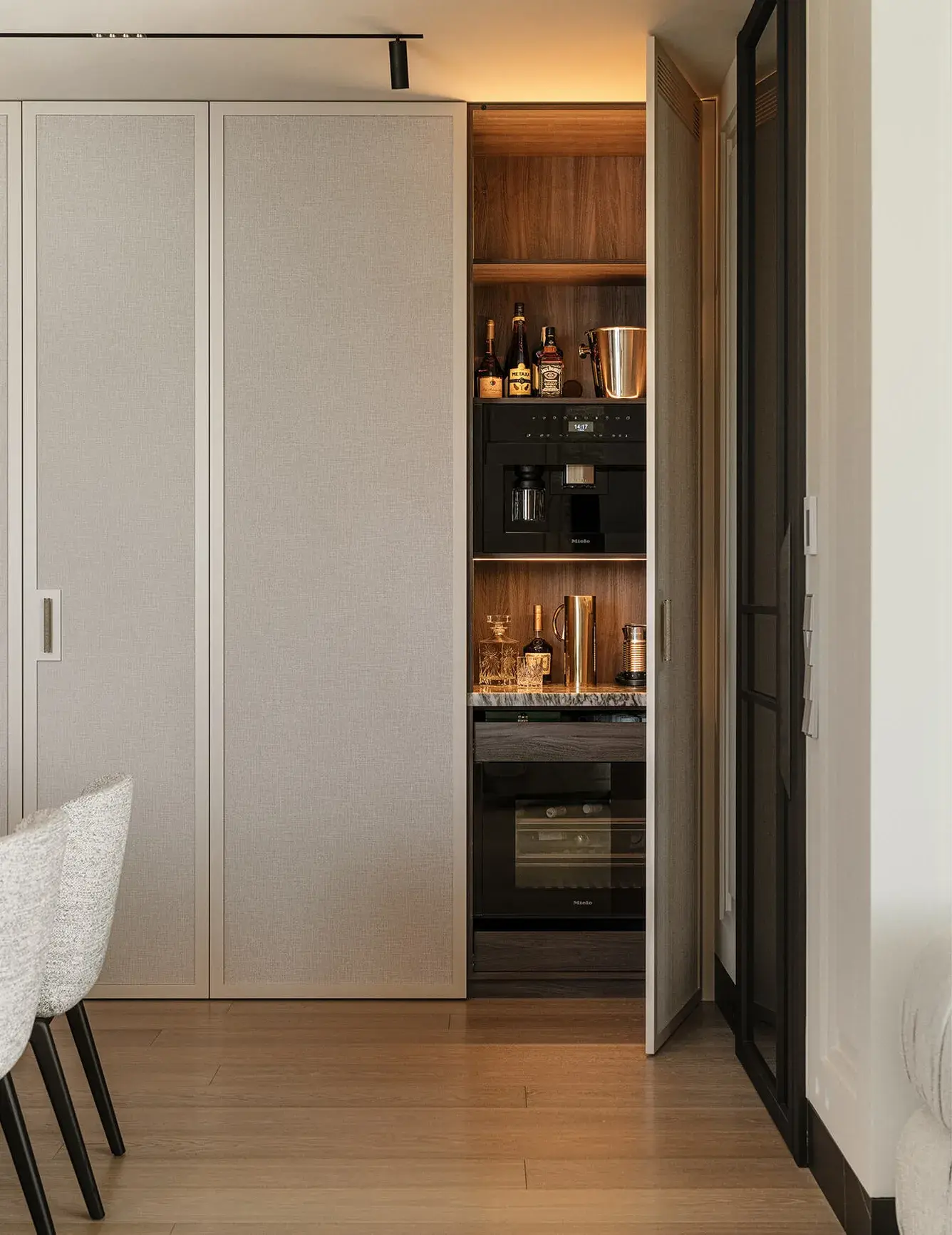 Detailní pohled na vestavěný bar a kávový koutek v bytě Iwicka, skrytý za světlými panelovými dveřmi, s interiérem z tmavého dřeva, profesionálním kávovarem a policemi na nápoje, vše doplněno dubovou podlahou Oakcent Premium v odstínu Milky.
