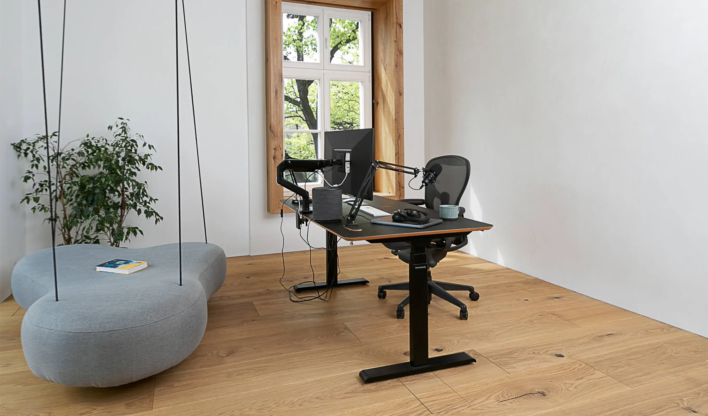 Moderní pracovní kout v apartmánu Moskal s minimalistickým černým stolem a technikou na dubové podlaze Oakcent Rustic, doplněný o designovou závěsnou pohovku ve tvaru oblázku a výhled skrze okno s masivním dřevěným ostěním.