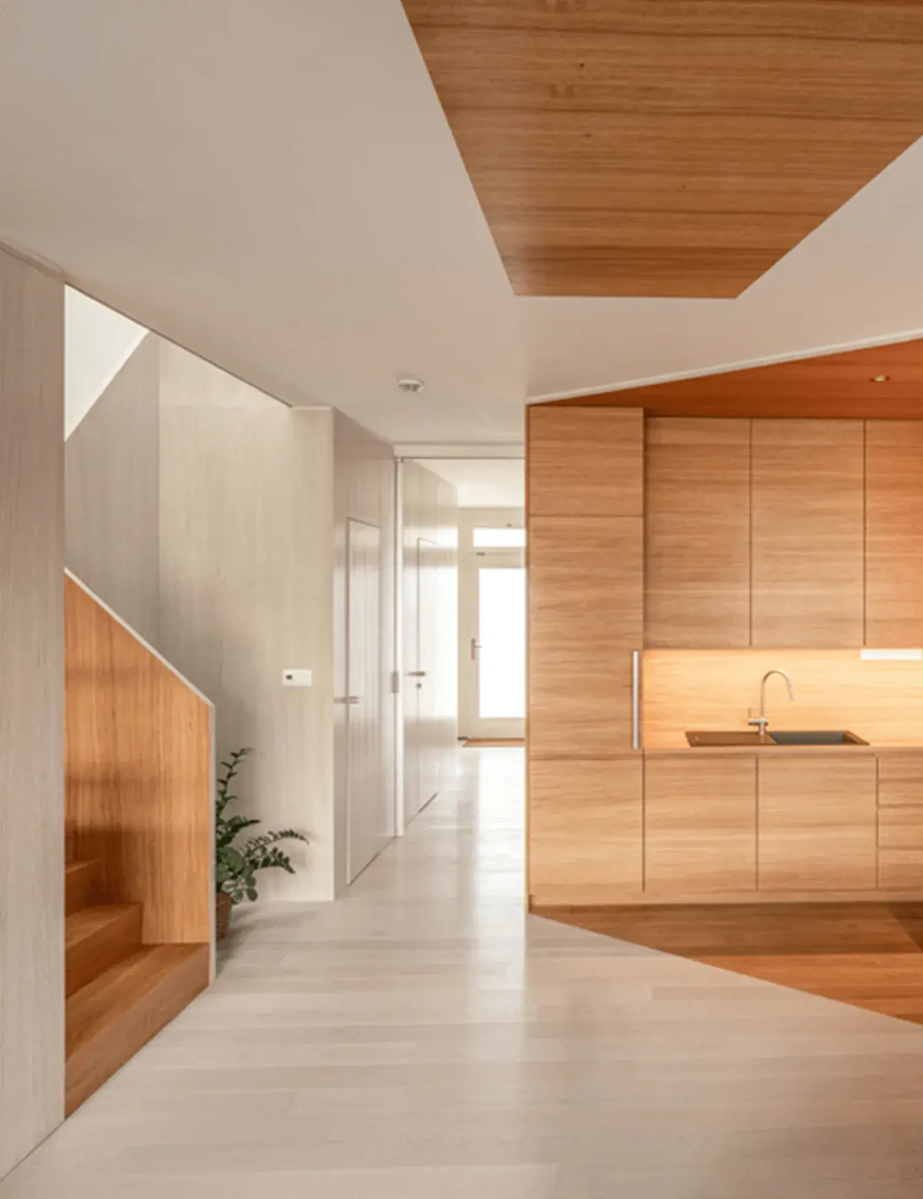 Pohled z chodby na propojený interiér rodinného domu Ke Káčátkům, kde světlá podlaha kontrastuje s dubovým schodištěm a vestavěnou kuchyňskou linkou s dřevěným podhledem, což vytváří ucelený a harmonický design celého podlaží.