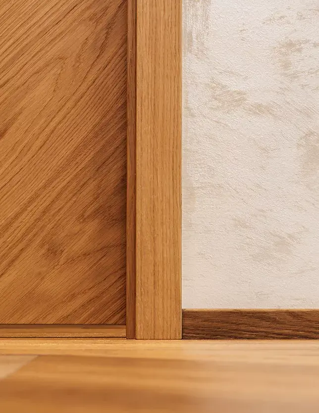 Detailní záběr na precizní napojení dubové podlahy, dveřní zárubně a obvodové lišty v showroomu Korbička, které ilustruje řemeslnou kvalitu instalace v kombinaci s dekorativní stěrkou na stěně.