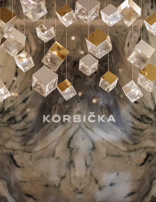 Detailní záběr na luxusní stropní svítidlo v showroomu Korbička, které se skládá z mnoha zavěšených skleněných a zlatých kostek, vytvářejících odlesky na pozadí mramorové stěny s logem šperkařství.