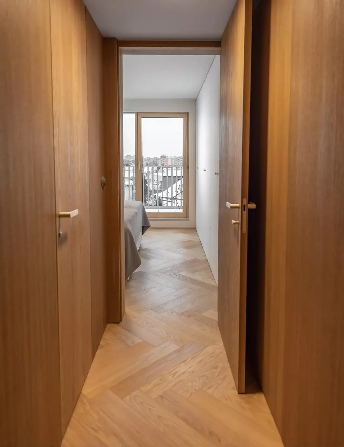 Pohled do ložnice v olomouckém bytě, kterému dominuje dubová podlaha Oakcent Sand položená do vzoru rybí kosti, plynule přecházející v celodřevěné obložení stěn a dveří, jež v kombinaci s bílou šatní skříní vytváří čistý a hřejivý interiér.