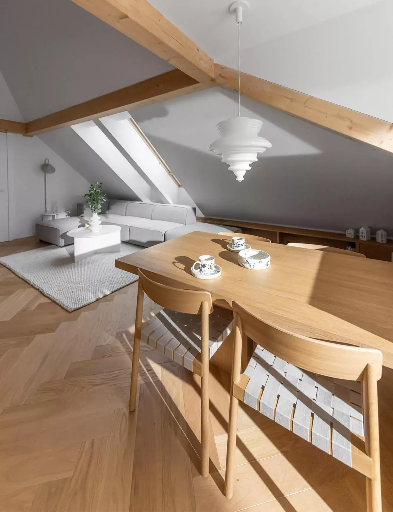 Prosvětlený interiér podkrovního bytu v Olomouci od Cojocarů Architekti, kde kartáčovaná dubová podlaha Oakcent Premium se vzorem rybí kosti (Herringbone) a přírodním olejem tvoří harmonický základ pro dřevěný jídelní stůl a odpočinkovou zónu s šedou pohovkou pod přiznanými trámy.