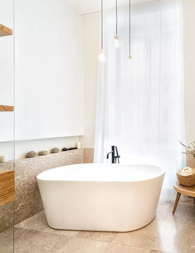 Interiér moderní světlé koupelny v brněnském bytě s volně stojící bílou vanou u okna s jemným závěsem, doplněnou mramorovým obkladem a designovým osvětlením.