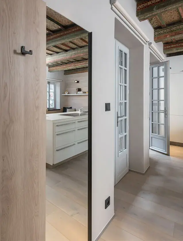 Průhled z chodby do moderní bílé kuchyně v historickém bytě na Malé Straně, zachycující detailní napojení světlé dubové podlahy Oakcent na bílé stěny a minimalistické interiérové dveře.