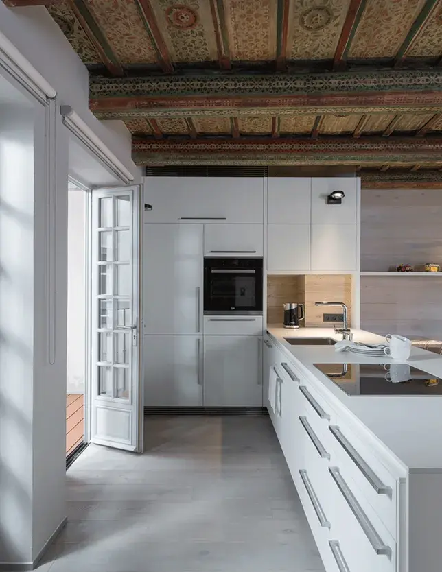 Celkový pohled na moderní bílou kuchyni s ostrůvkem v historickém bytě na Malé Straně, zdůrazňující soulad světlé dubové podlahy Oakcent z kolekce Ivory a restaurovaného renesančního záklopového stropu s ornamentální malbou.