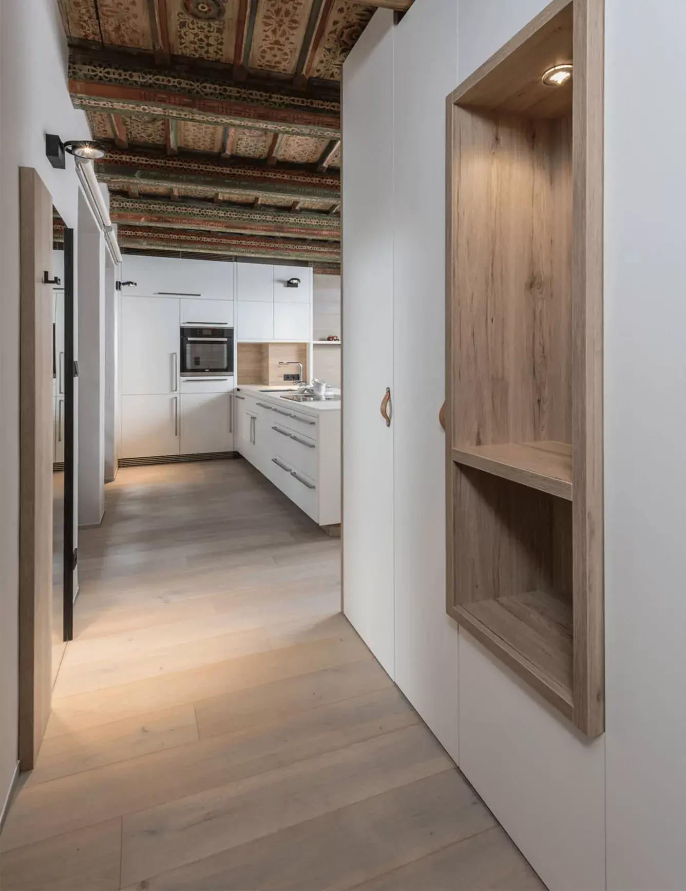 Pohled do chodby historického bytu na Malé Straně s vestavnou dřevěnou policí, bílými skříněmi a světlou dubovou podlahou Oakcent, která plynule přechází do moderní bílé kuchyně.