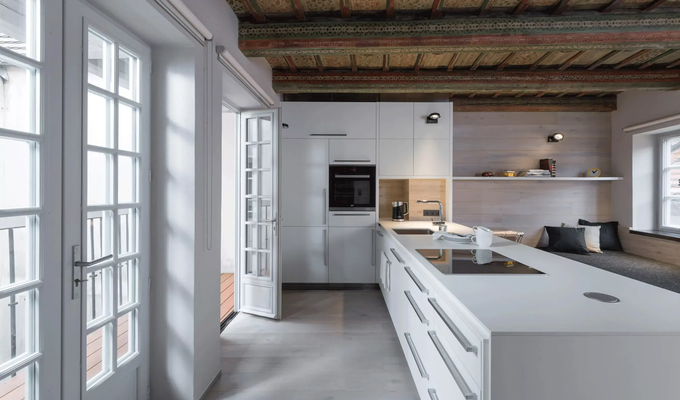 Alt text: Pohled do moderní bílé kuchyně s ostrůvkem v historickém bytě na Malé Straně, zachycující kontrast minimalistického designu, původního malovaného trámového stropu a světlé dubové podlahy Oakcent.