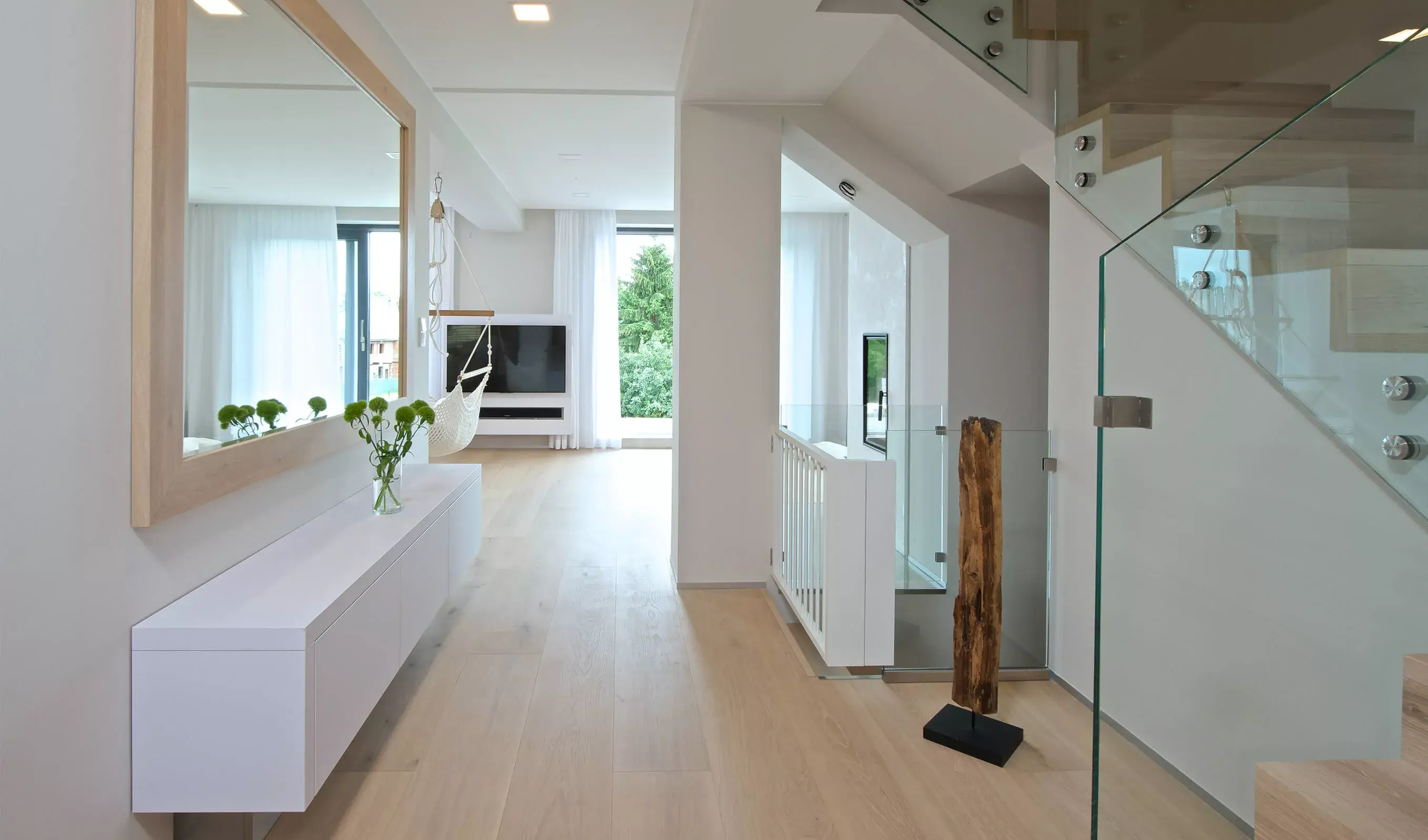 Široký pohled na prosvětlenou vstupní halu soukromého domu, kde dubová podlaha Oakcent Rustic v odstínu Light White plynule přechází do obývacího prostoru a harmonizuje s bílým minimalistickým nábytkem a velkým zrcadlem v dřevěném rámu.