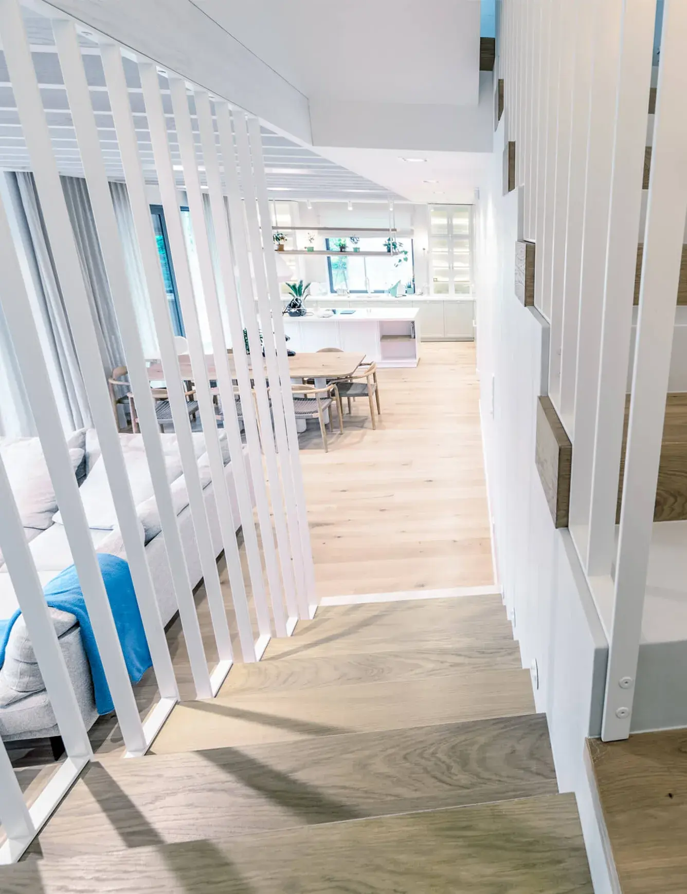 Pohled ze schodiště do otevřeného přízemí soukromého domu v Krakově, kde dominují světlé tóny, bílé vertikální lamely zábradlí a kouřovaná dubová podlaha Oakcent Rustic v přírodním odstínu Pure.