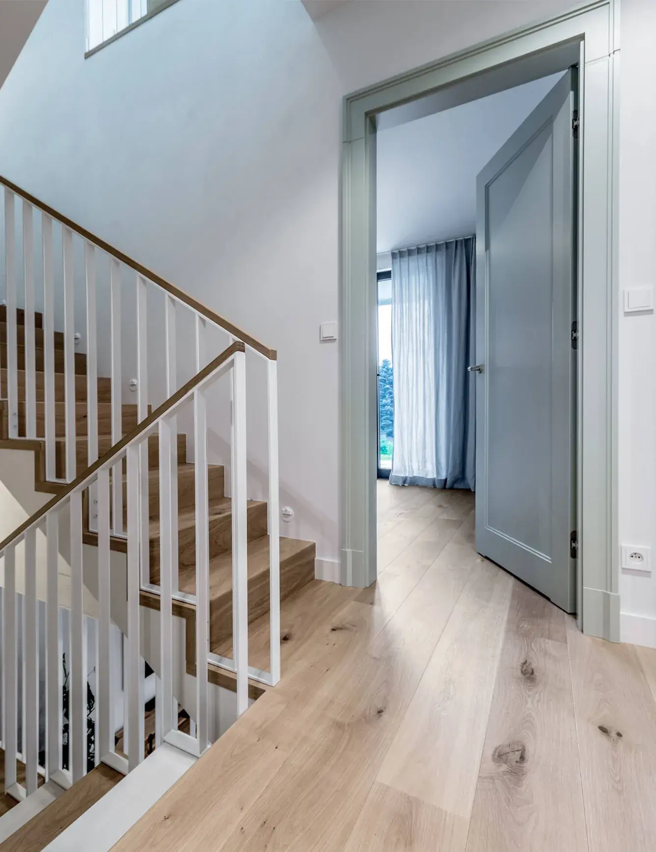 Detailní pohled na horní chodbu krakovského domu Motyla, kde kouřovaná dubová prkna Oakcent Rustic v přírodním odstínu Pure plynule navazují na dřevěné schodiště s bílým lamelovým zábradlím a minimalistické světle šedé interiérové dveře.