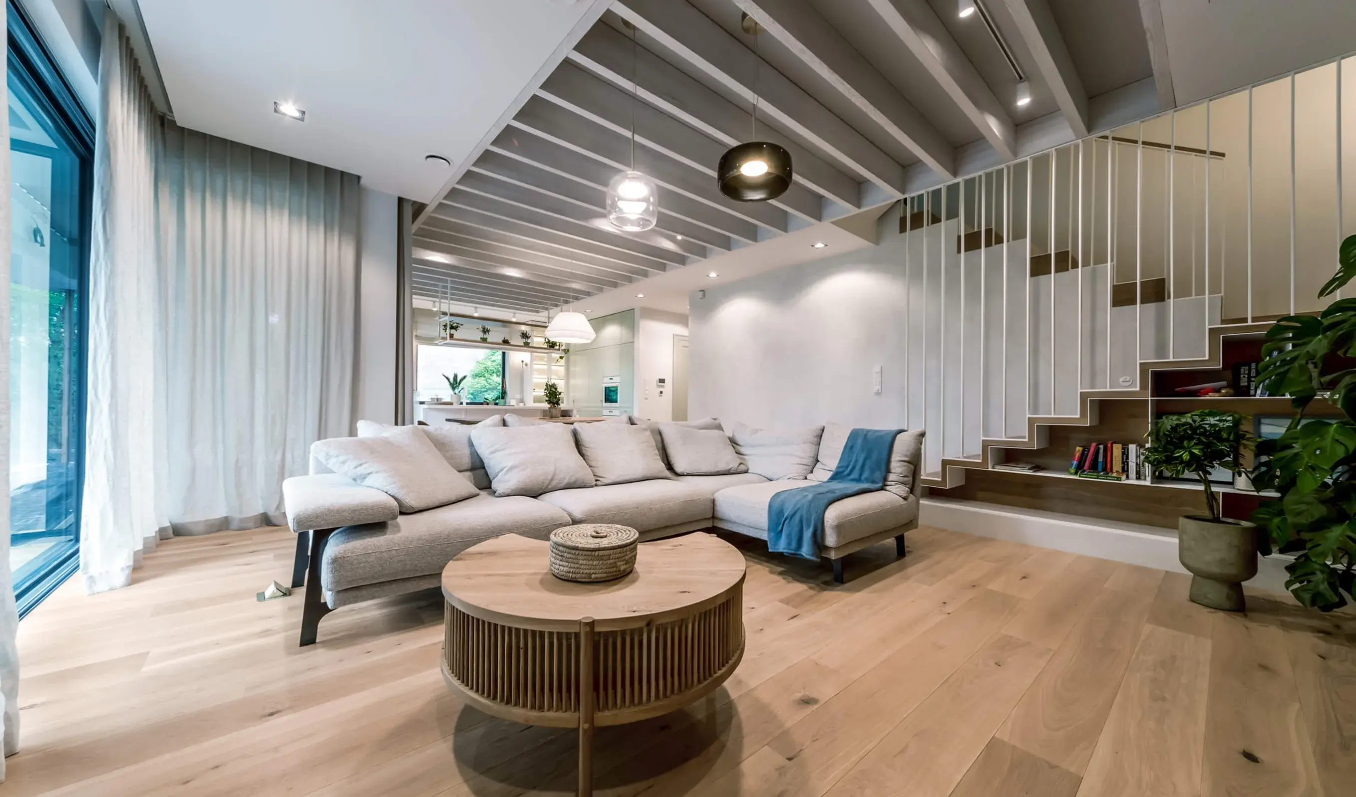 Široký pohled do moderního obývacího pokoje v krakovském domě Motyla, kde kouřovaná dubová podlaha Oakcent Rustic v přírodním odstínu Pure vkusně doplňuje minimalistickou bílou barvu stěn, jemné textury šedé sedací soupravy a originální schodiště s lamelovým zábradlí.