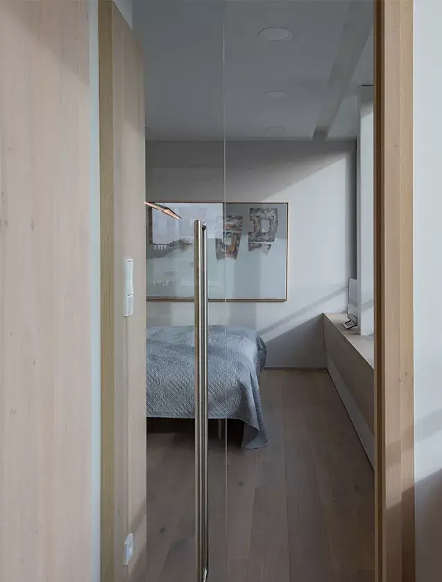 Pohled skrze skleněné dveře do minimalistické ložnice v pražském střešním bytě od ID_arch, kde světlé tóny stěn a vestavěného nábytku doplňuje kartáčovaná, lehce kouřovaná dubová podlaha Oakcent ošetřená Milky olejem.