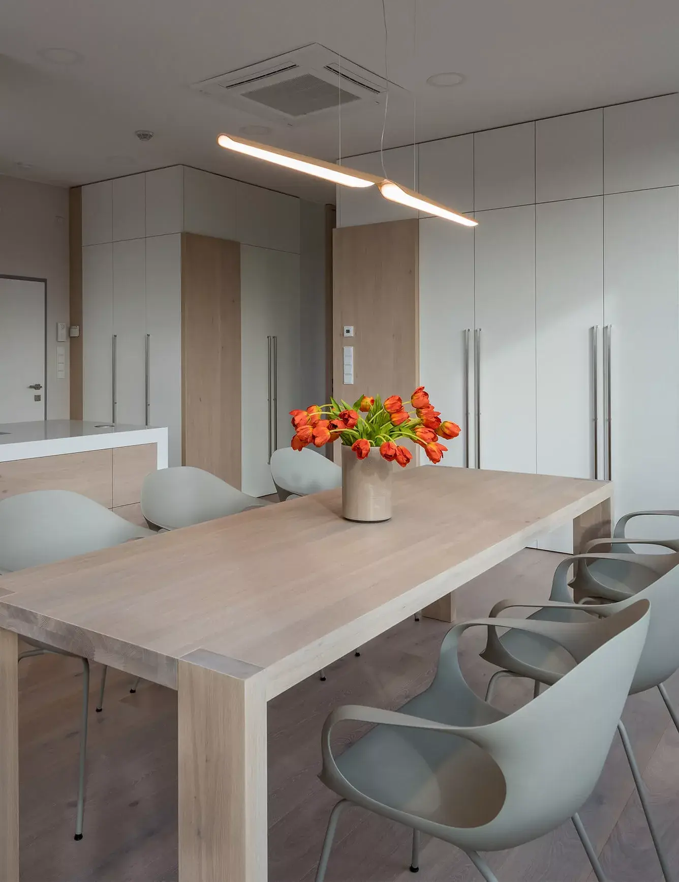 Detail jídelního koutu v pražském střešním bytě od ID_arch, kterému dominuje masivní dřevěný stůl ladící s kartáčovanou dubovou podlahou Oakcent v odstínu Milky, doplněný o moderní šedé židle, vázu s oranžovými tulipány a minimalistické lineární svítidlo.