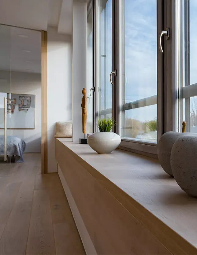 Detailní pohled na nízký dubový parapet, který se táhne podél velkoformátových oken střešního bytu a barevně i materiálově navazuje na podlahu Oakcent Sand, čímž dotváří klidný a sjednocený charakter celého interiéru.