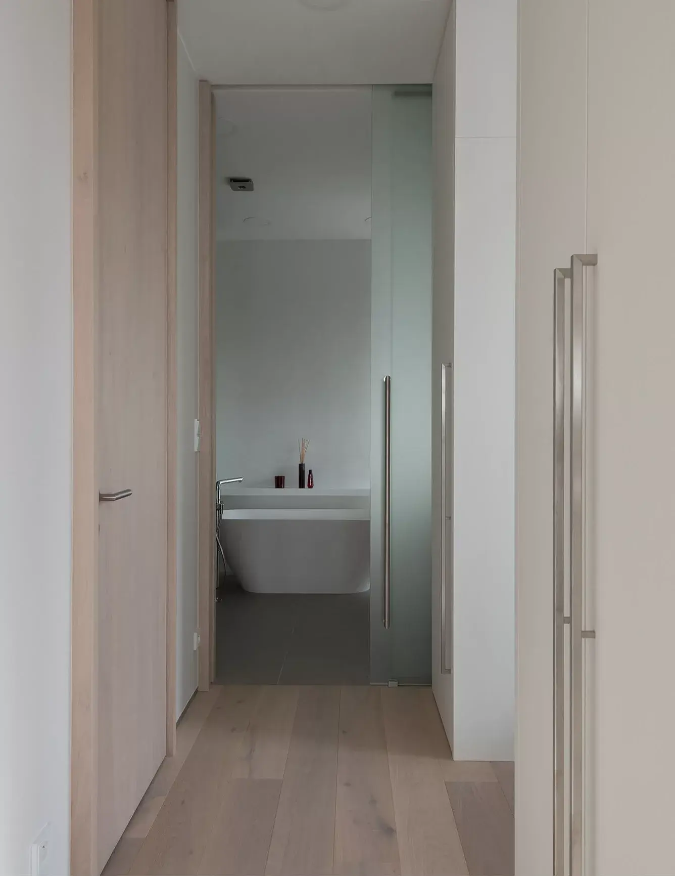 Průhled z chodby se světlou dubovou podlahou Oakcent Milky do minimalistické koupelny pražského střešního bytu, kde se za posuvnými skleněnými dveřmi nachází volně stojící bílá vana u čisté bílé stěny.