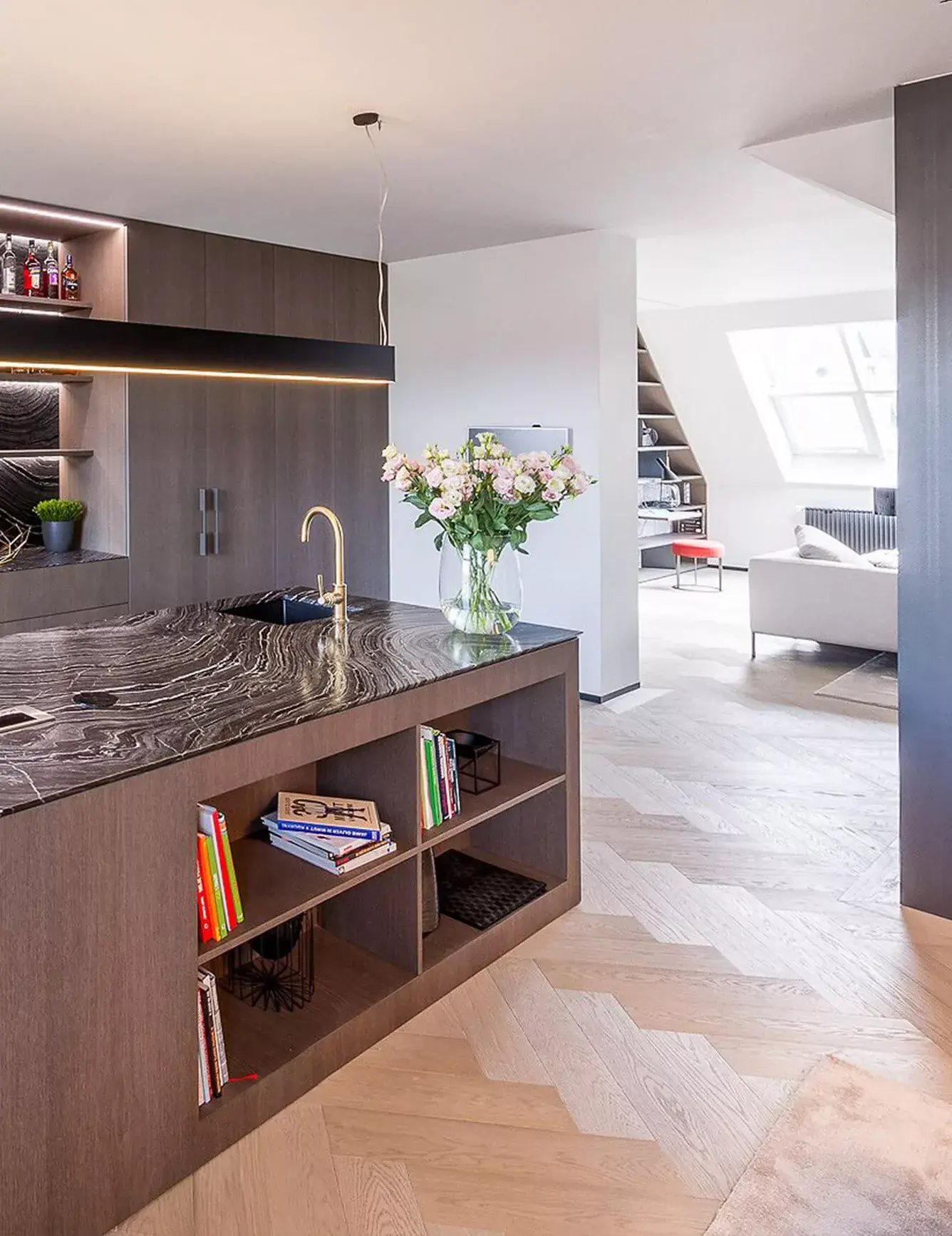 Detailní pohled na moderní kuchyni s mramorovým ostrůvkem v podkrovním pražském apartmánu, kde dubová podlaha Oakcent Premium Herringbone v odstínu Quartzgrau plynule prochází do prosvětleného obývacího pokoje s vestavěnou knihovnou.