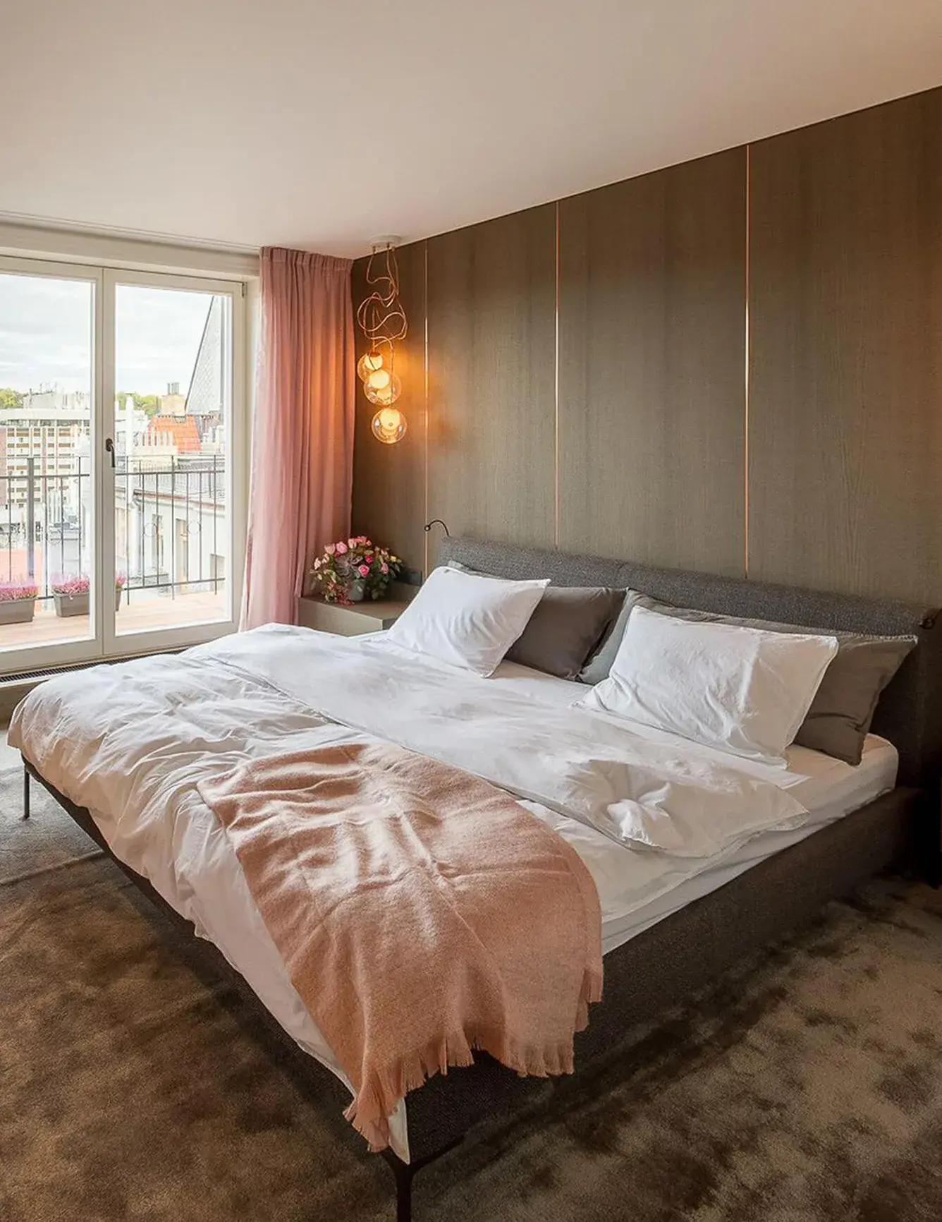 Detailní pohled na elegantní ložnici ve staroměstském apartmánu, kde tmavé dřevěné obložení stěny s jemnými vertikálními lištami vytváří luxusní pozadí pro šedé čalouněné lůžko, doplněné o designové skleněné svítidlo a výhled na pražské střechy.