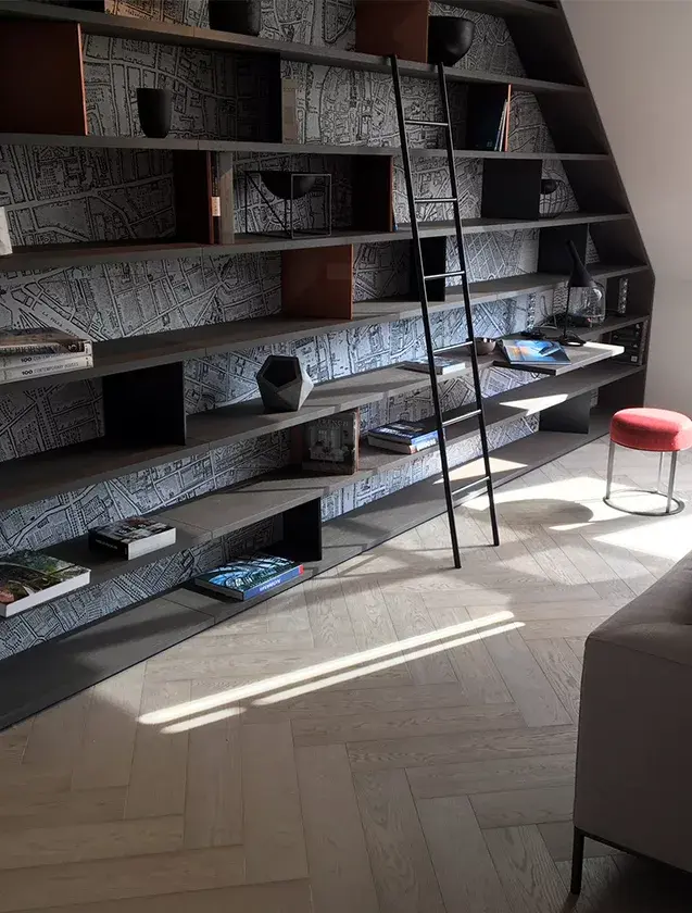 Detailní pohled na dubovou podlahu Oakcent Premium ve vzoru Herringbone (stromeček) v šedém odstínu Quartzgrau, která podtrhuje moderní charakter obývacího pokoje s rozměrnou vestavěnou knihovnou a elegantním světlým nábytkem.